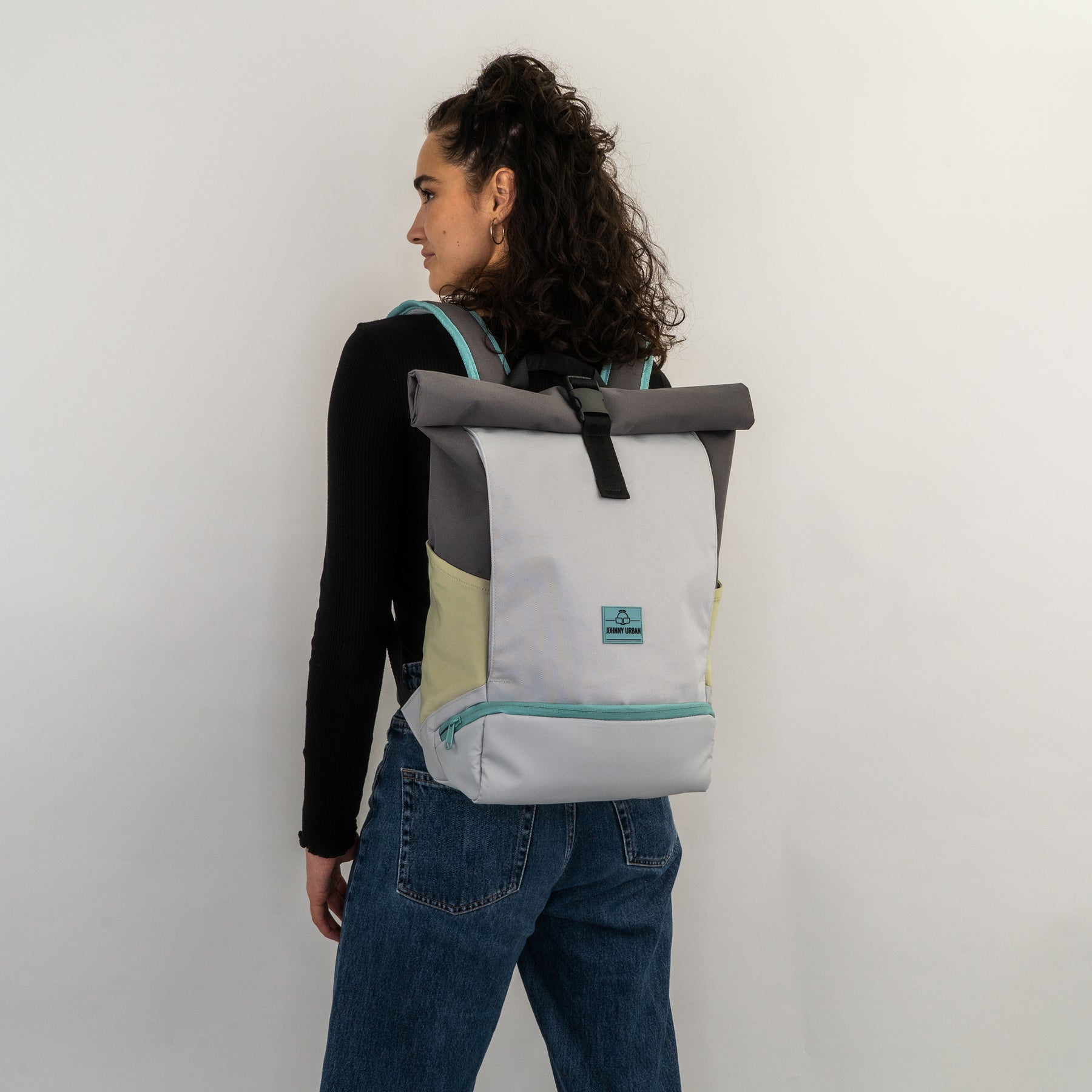 Frau mit dunklen lockigen Haaren trägt Rolltop Rucksack 'Allen Medium' in hellgrau, grau und beige mit türkisem Reißverschluss und Logo