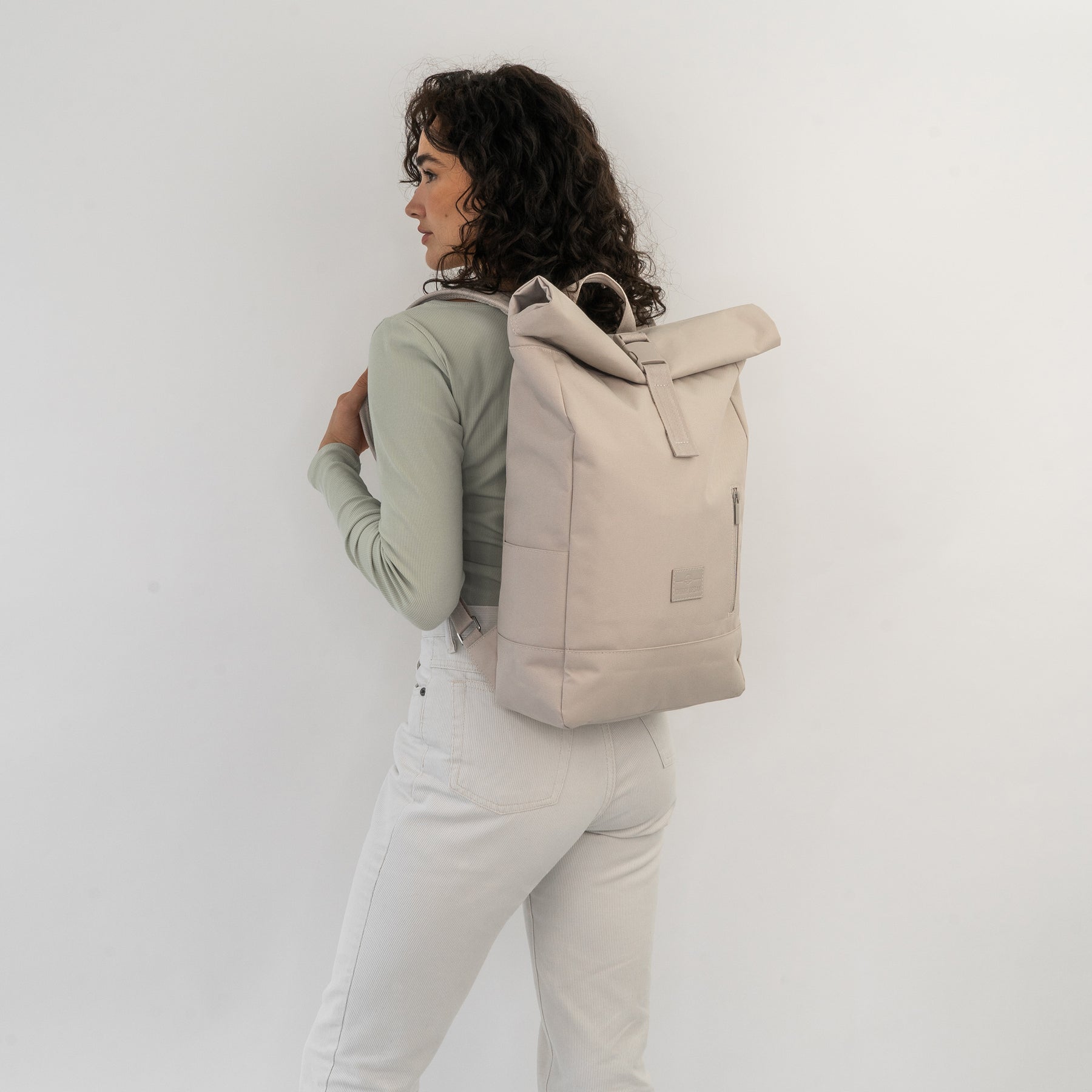 Frau mit lockigen Haaren trägt hellbeigen Rolltop Rucksack 'Robin Medium' vor weißem Hintergrund