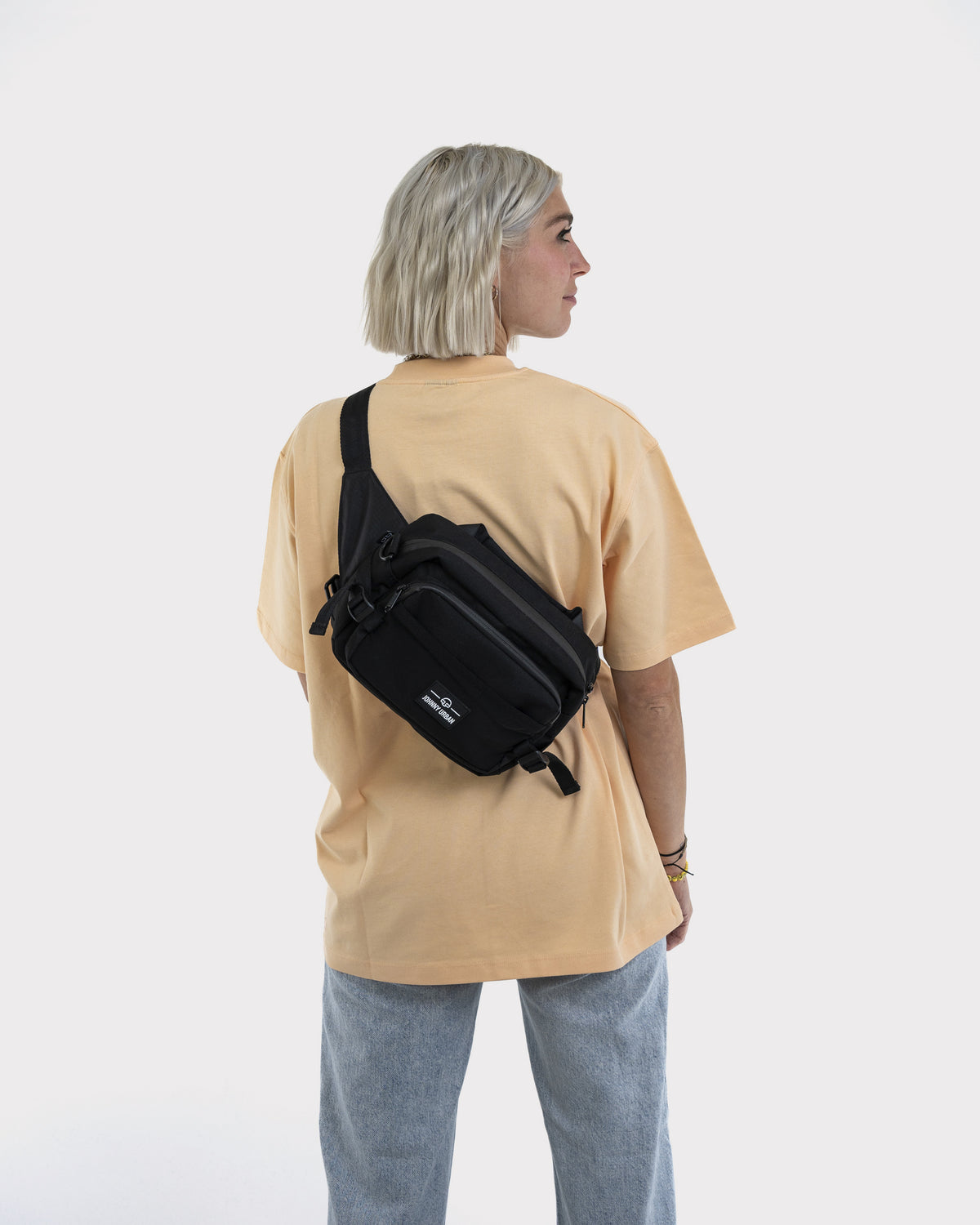 Frau trägt schwarze Johnny Urban Bauchtasche 'Travis' diagonal am Rücken mit beiger T-Shirt und blauen Jeans