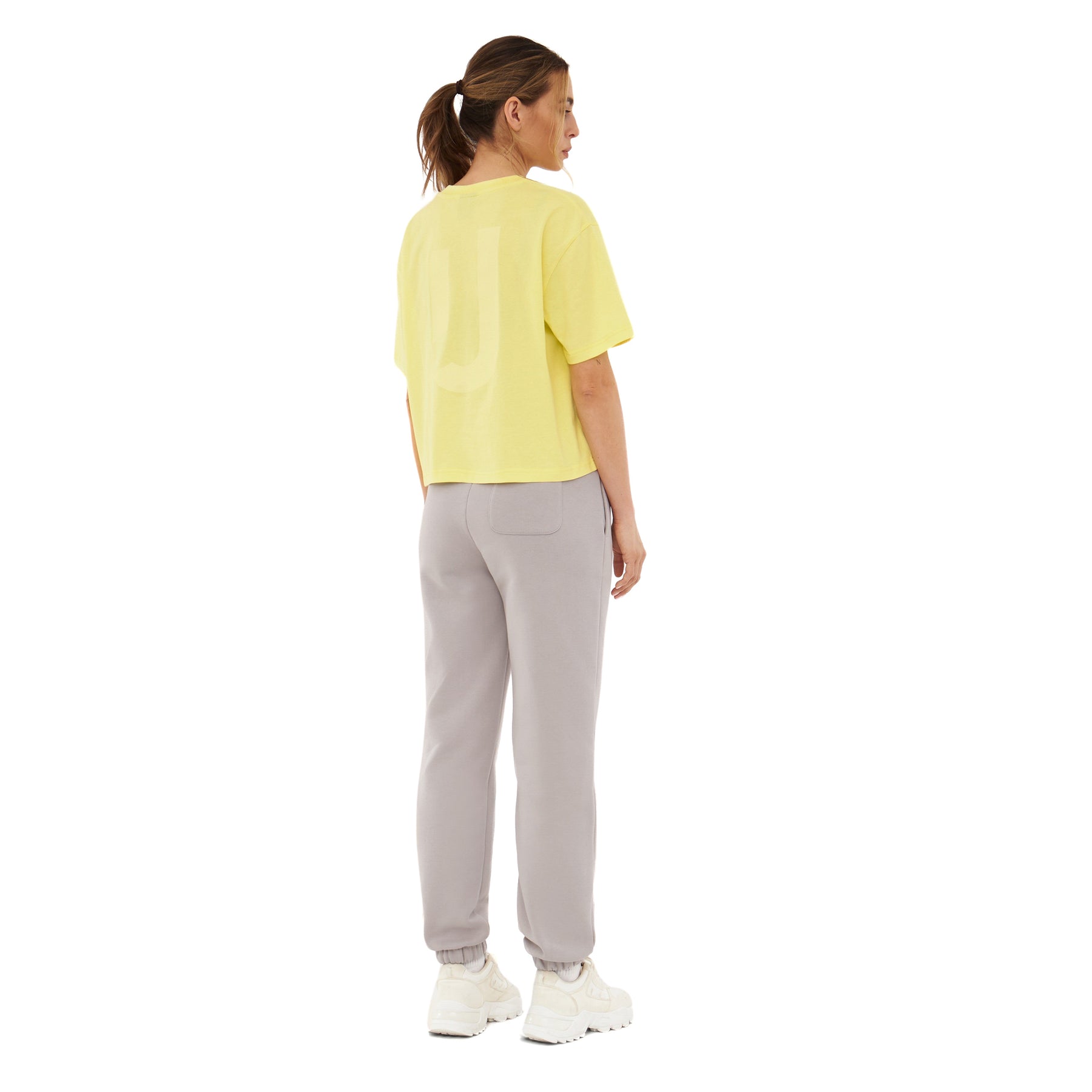 Frau von hinten in gelbem Cropped T-Shirt 'Zoey JU' und hellgrauen Jogginghosen mit weißen Sneakern