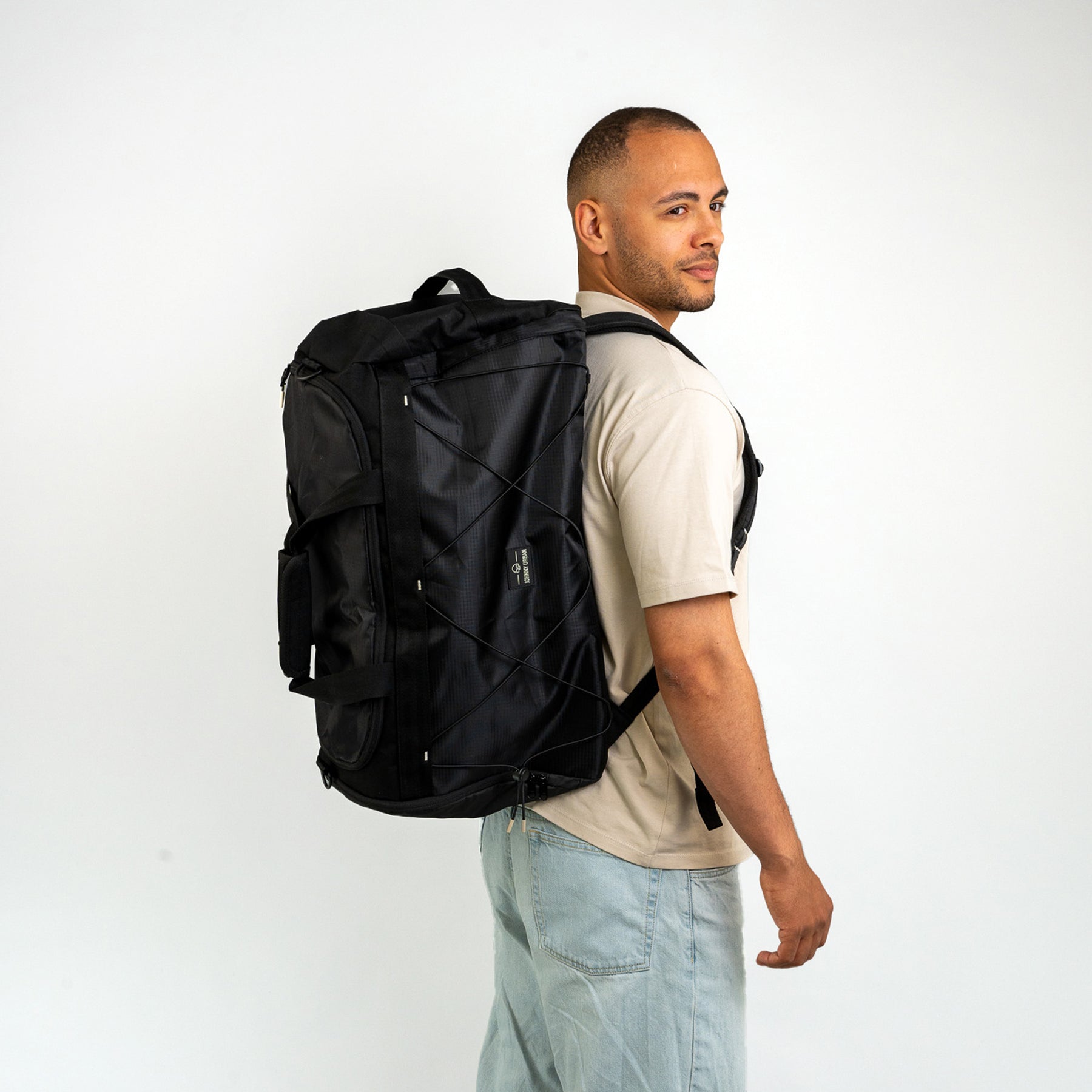 Mann trägt schwarzen Duffle Bag 'Shawn Large' auf Rücken, beige T-Shirt, helle Jeans