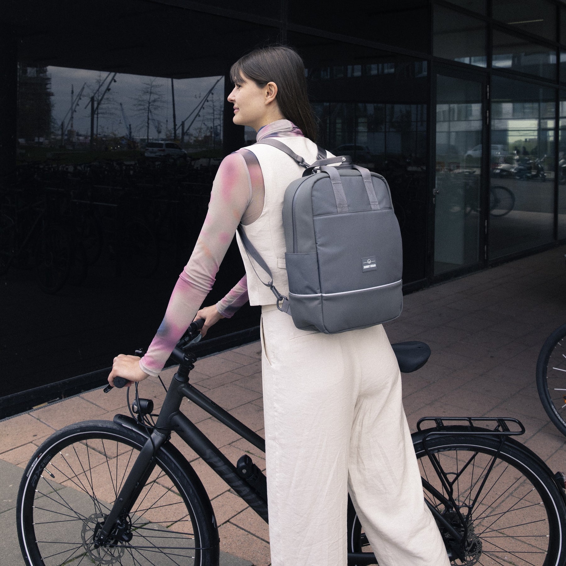 Frau mit grauem Johnny Urban Rucksack 'Jona Medium' lehnt an schwarzem Fahrrad