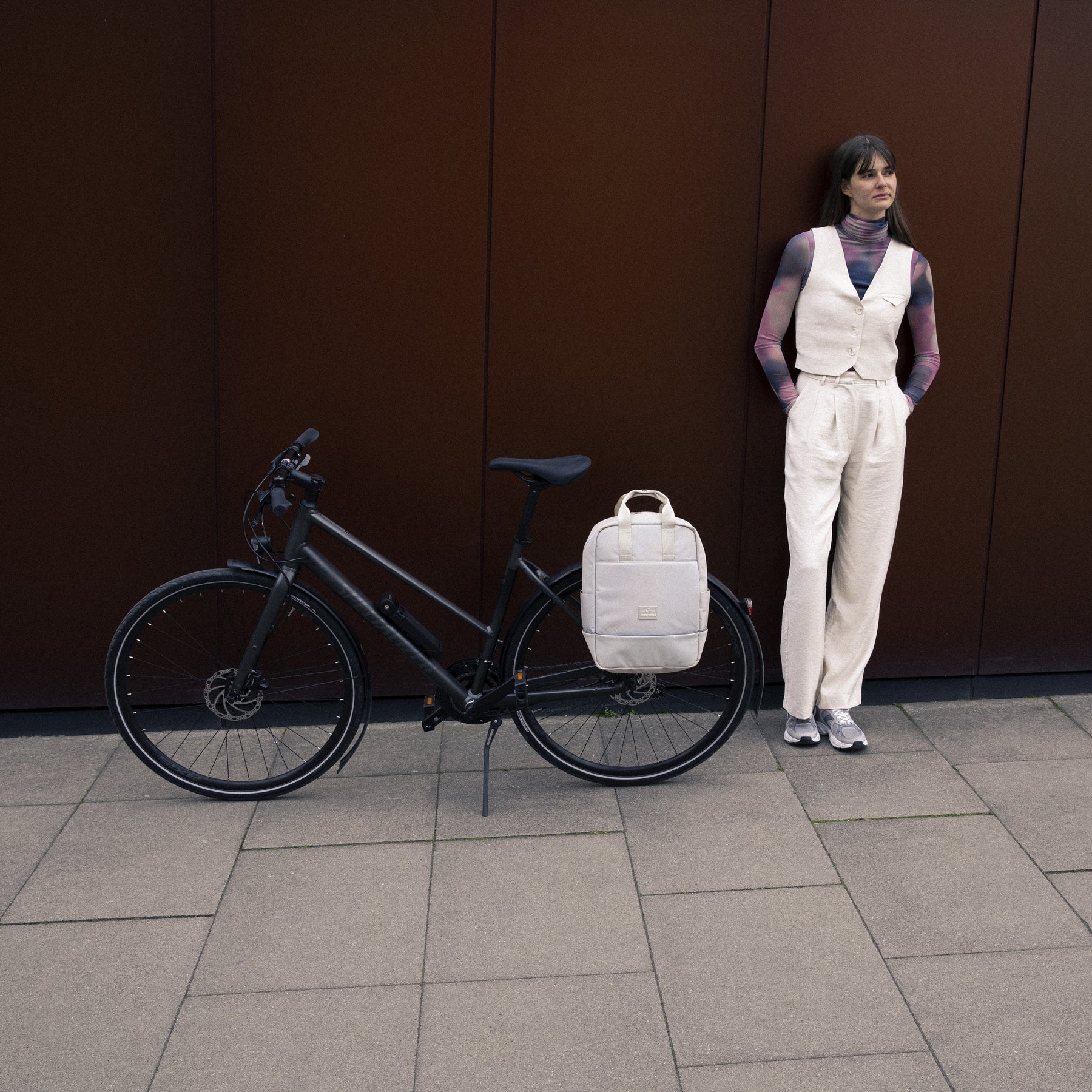 Fahrrad mit beigefarbenem Johnny Urban Rucksack 'Jona Medium' am Gepäckträger, daneben steht eine Frau in heller Hose und Weste