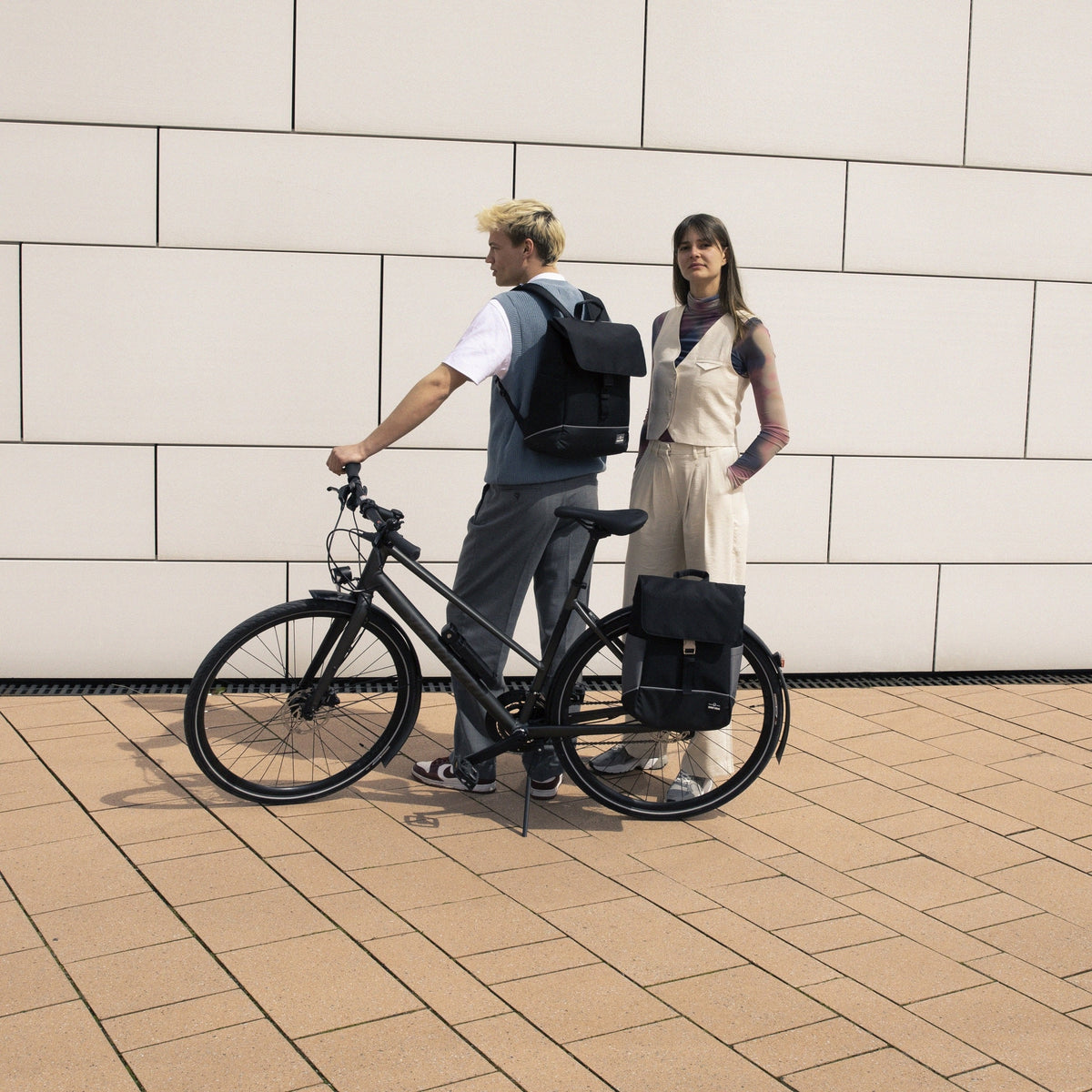 Mann mit schwarzem Rucksack 'Mika' auf einem schwarzen Fahrrad, Frau neben ihm mit schwarzem Rucksack 'Mika' neben Fahrrad