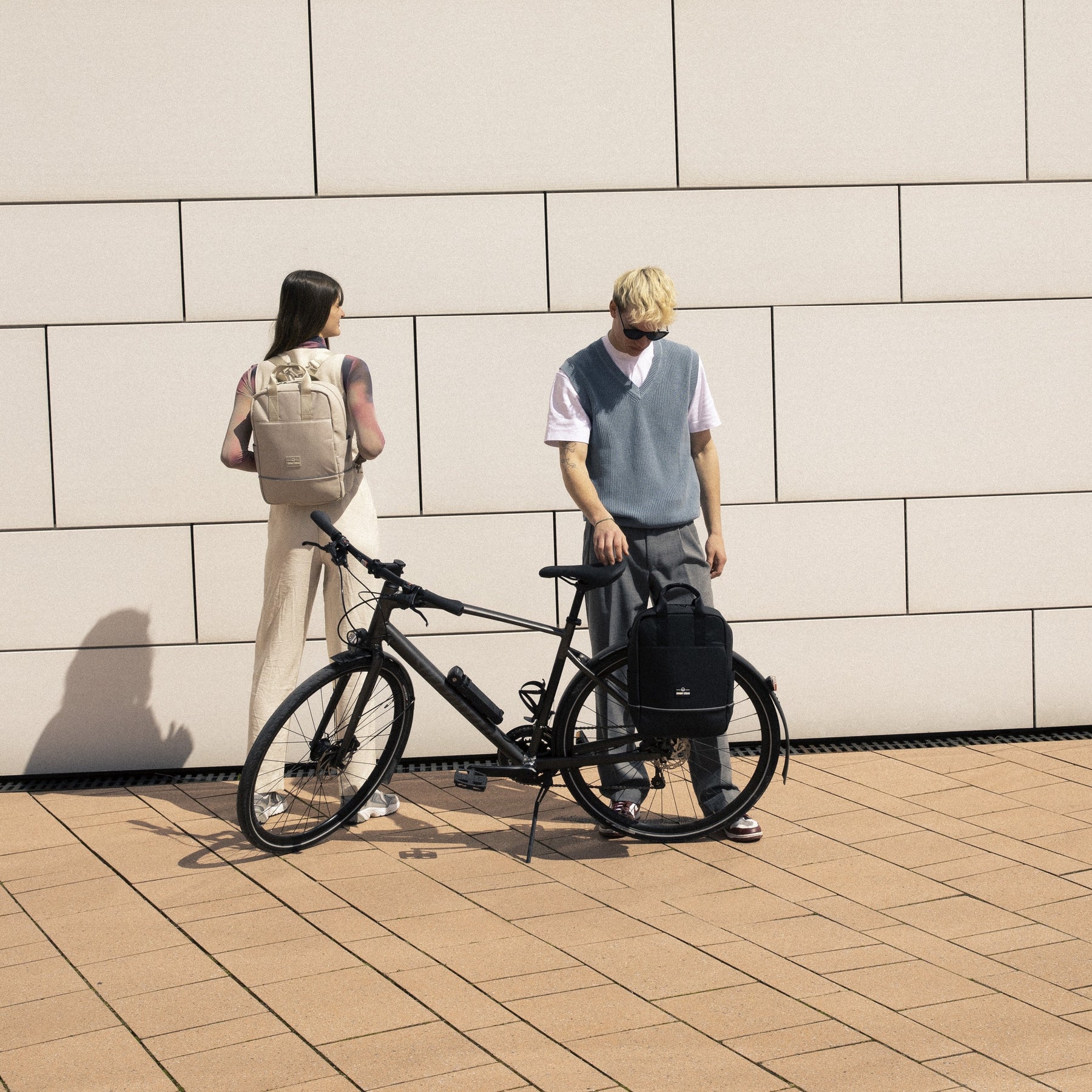 Frau mit beigem Johnny Urban Rucksack 'Jona Medium' und Mann mit schwarzem Rucksack am Fahrrad vor hellen Fliesenwand
