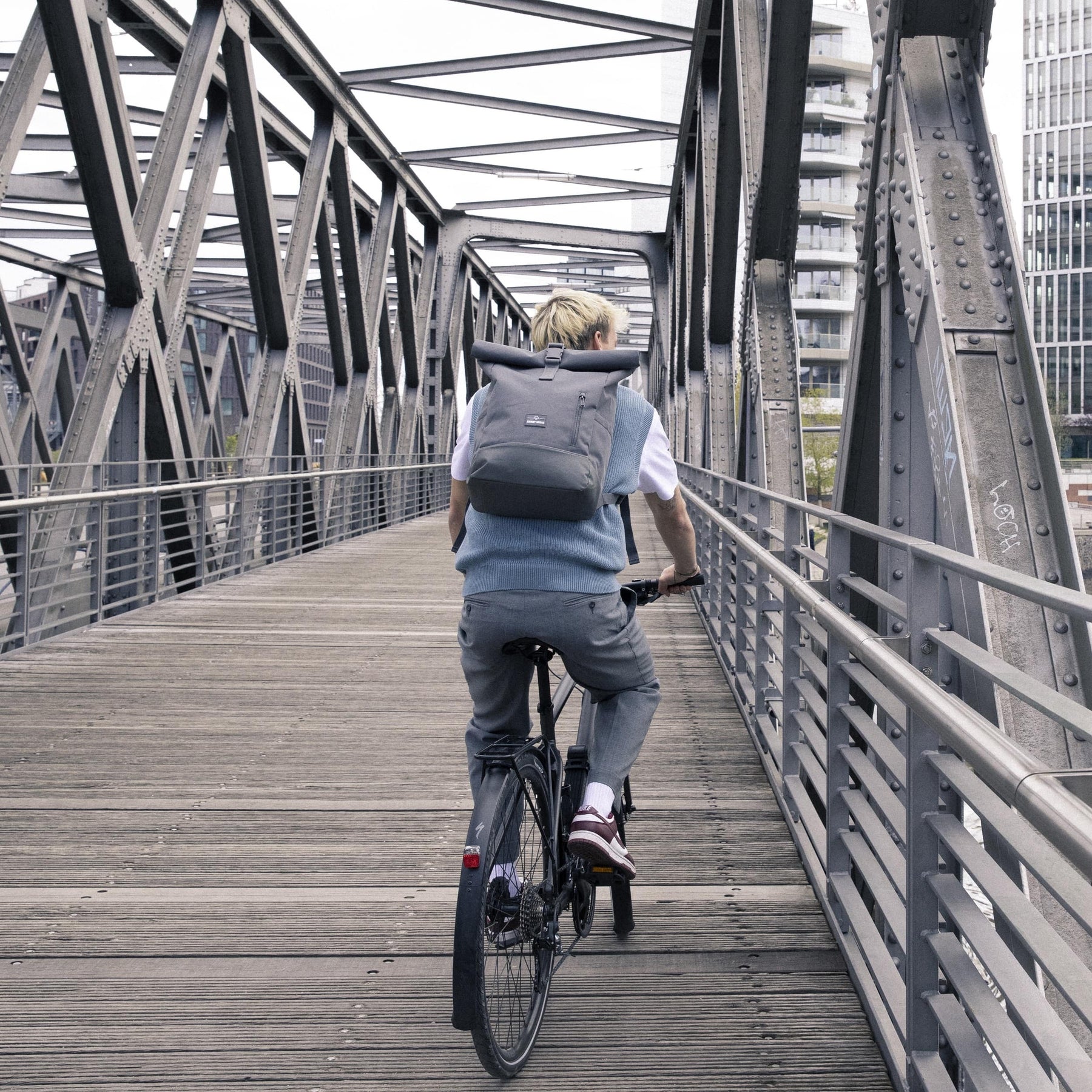Mann fährt mit Rolltop Rucksack 'Robin Medium' auf einem Fahrrad über eine stählerne Fußgängerbrücke in der Stadt
