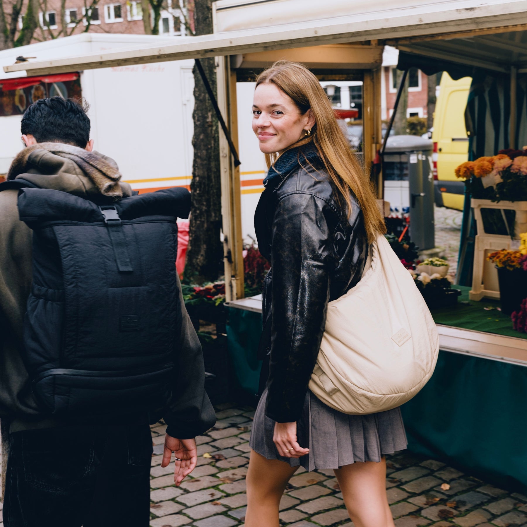 Frau trägt beige Half Moon Bag 'Kaia XL' von Johnny Urban, neben ihr Mann mit großem schwarzem Rucksack, Marktstand im Hintergrund