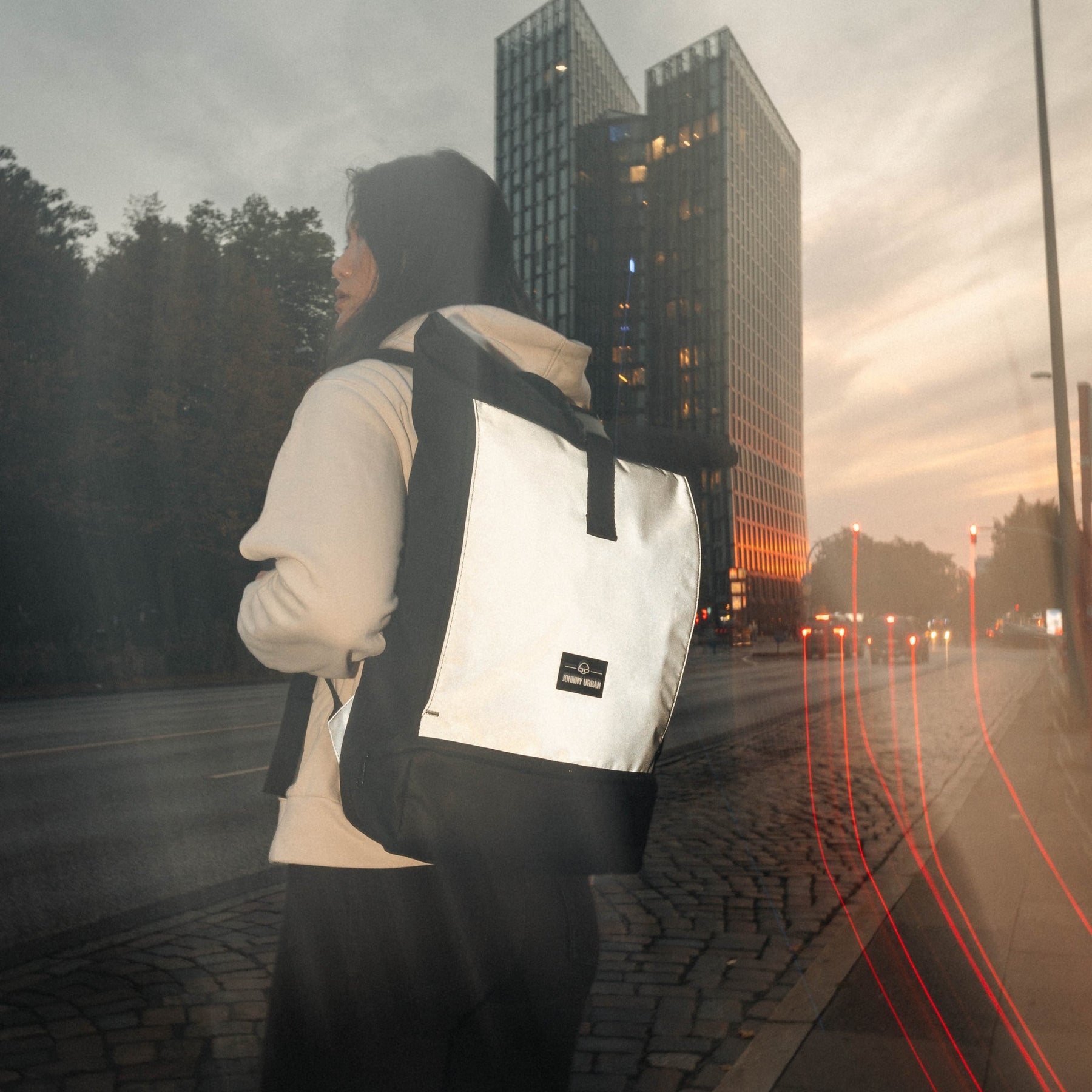 Frau trägt reflektierenden Rolltop Rucksack 'Allen Large' in urbaner Umgebung bei Dämmerung