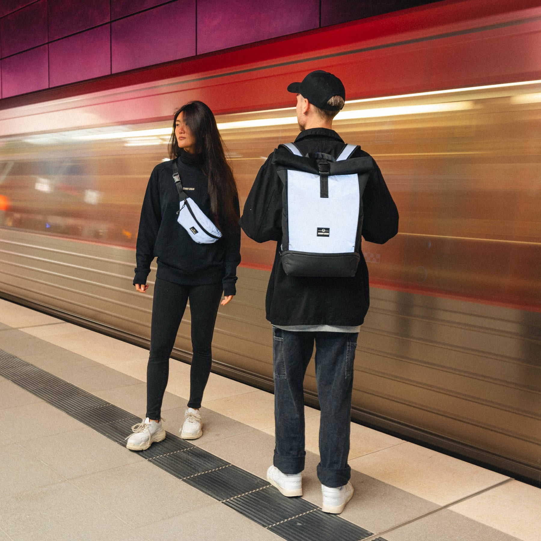 Zwei Personen an einer U-Bahn-Station, einer trägt den reflektierenden Rolltop Rucksack ‚Allen Medium‘ von Johnny Urban, der andere eine reflektierende Schultertasche