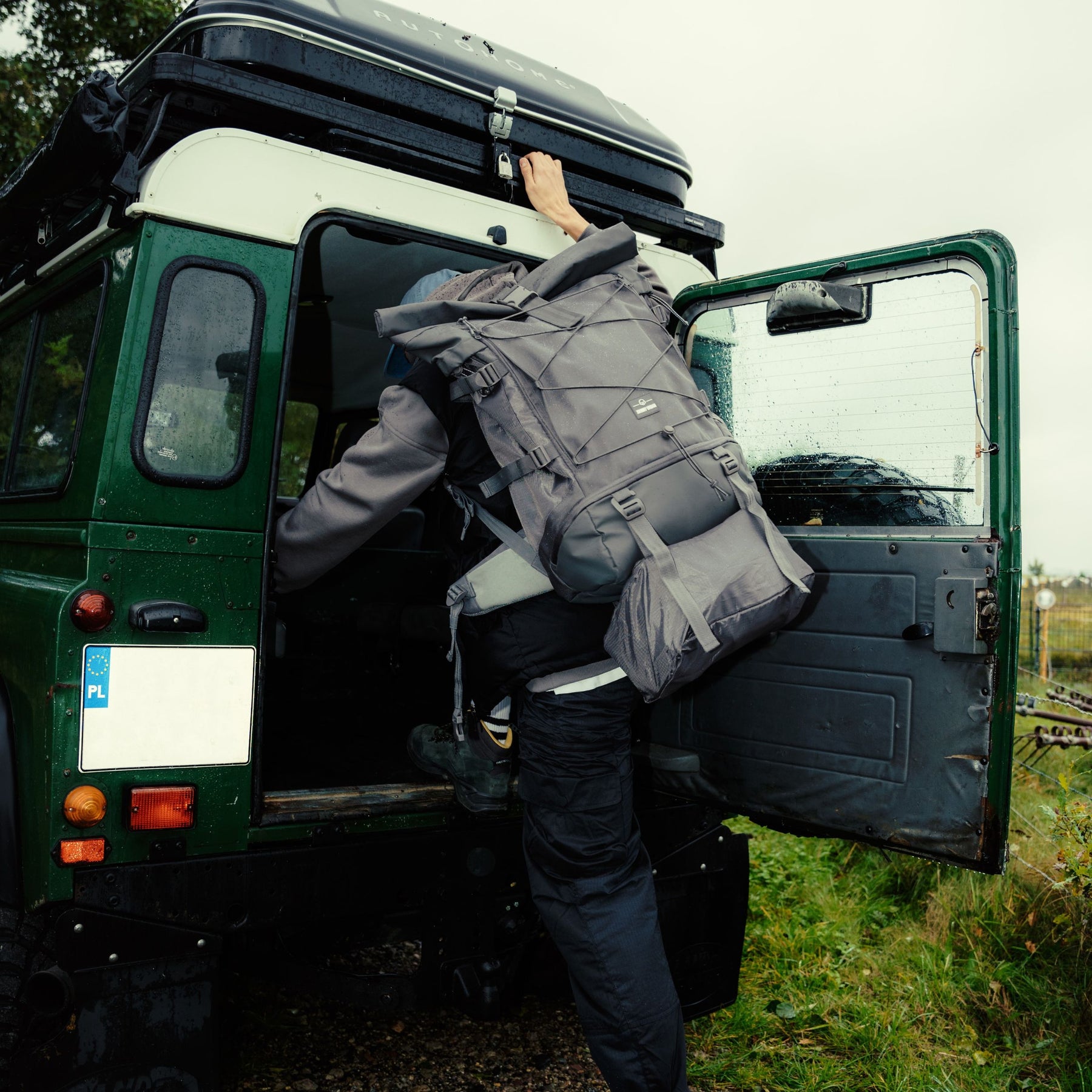 Person klettert mit großem grauem Johnny Urban Allen XL Travel Reiserucksack in grünes Geländefahrzeug