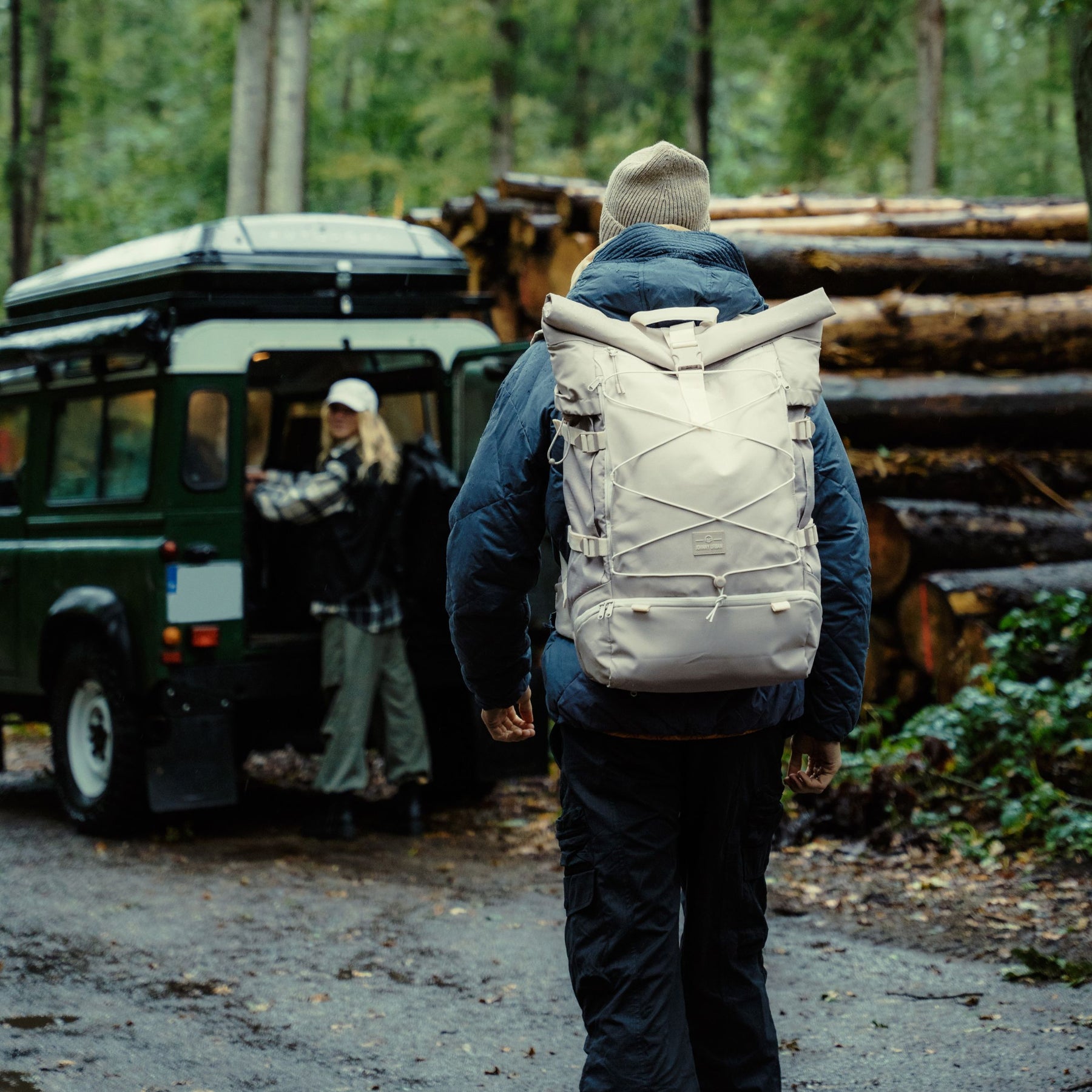 Mann trägt beigen Johnny Urban Reiserucksack 'Allen XL Travel' im Wald, Frau steht neben grünem Geländewagen