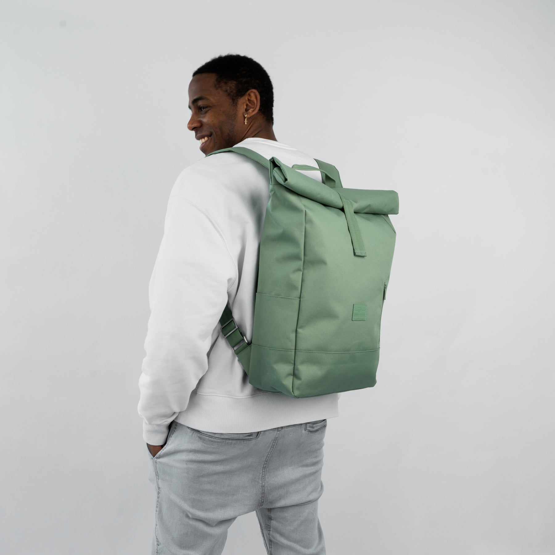 Mann trägt grünen Rolltop-Rucksack 'Robin Large' von Johnny Urban, kombiniert mit weißem Pullover und hellen Jeans