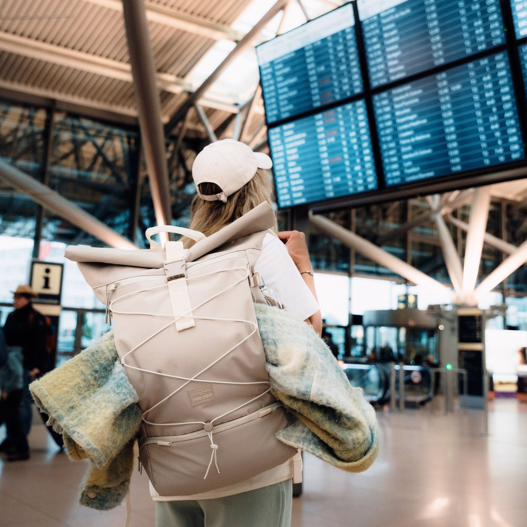 Frau an einem Flughafen mit beigefarbenem Rolltop-Reiserucksack ‚Allen Large Travel‘ von Johnny Urban auf dem Rücken