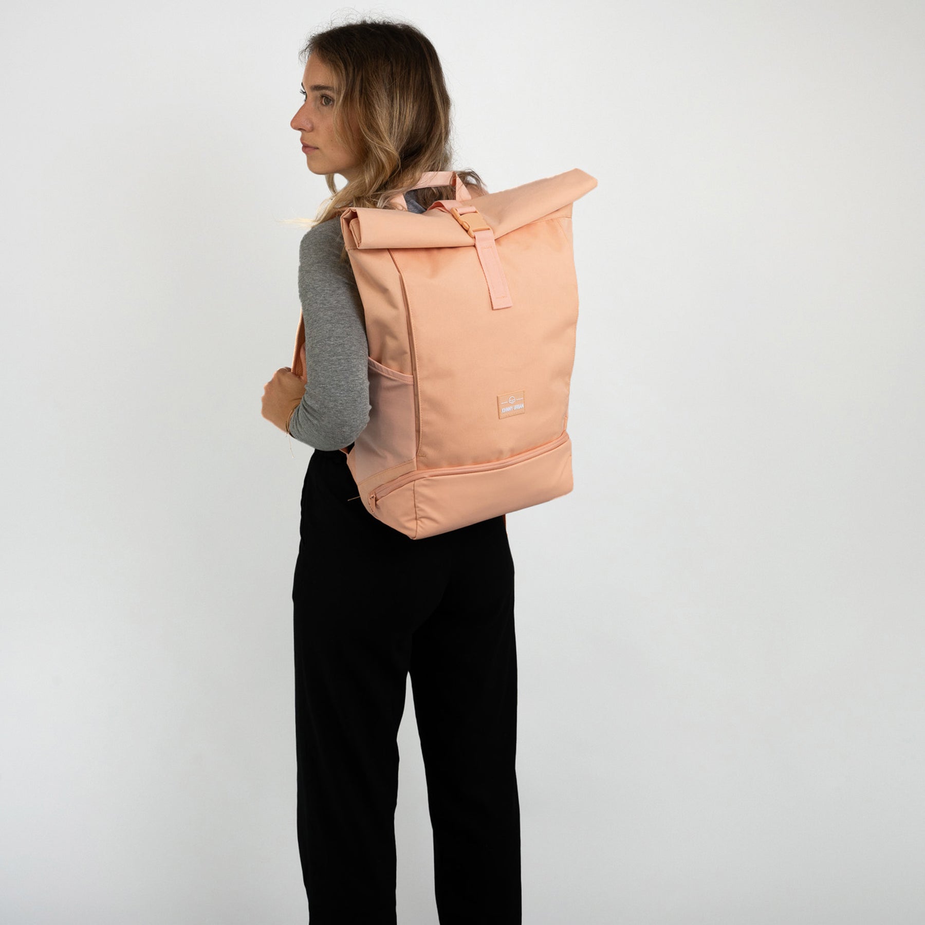 Frau trägt Rolltop Rucksack 'Allen Large' in Pfirsichfarbe mit grauem Oberteil und schwarzer Hose