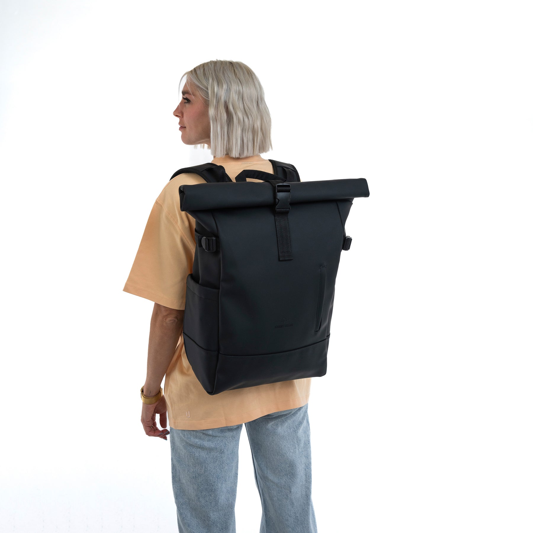 Frau mit kurzem blondem Haar trägt schwarzen Rolltop Rucksack 'Harvey Large' von hinten in lässiger Kleidung