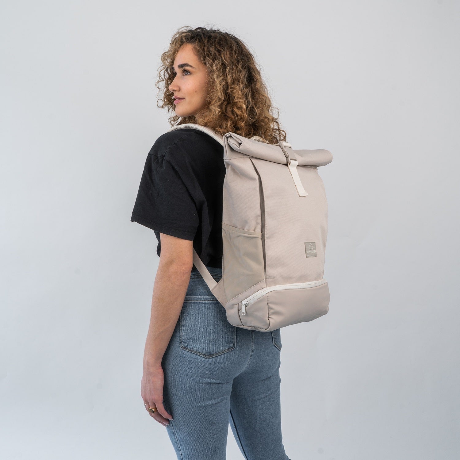 Frau mit lockigen Haaren trägt beigen Rolltop Rucksack 'Allen Medium' von Johnny Urban, schwarze T-Shirt, blaue Jeans
