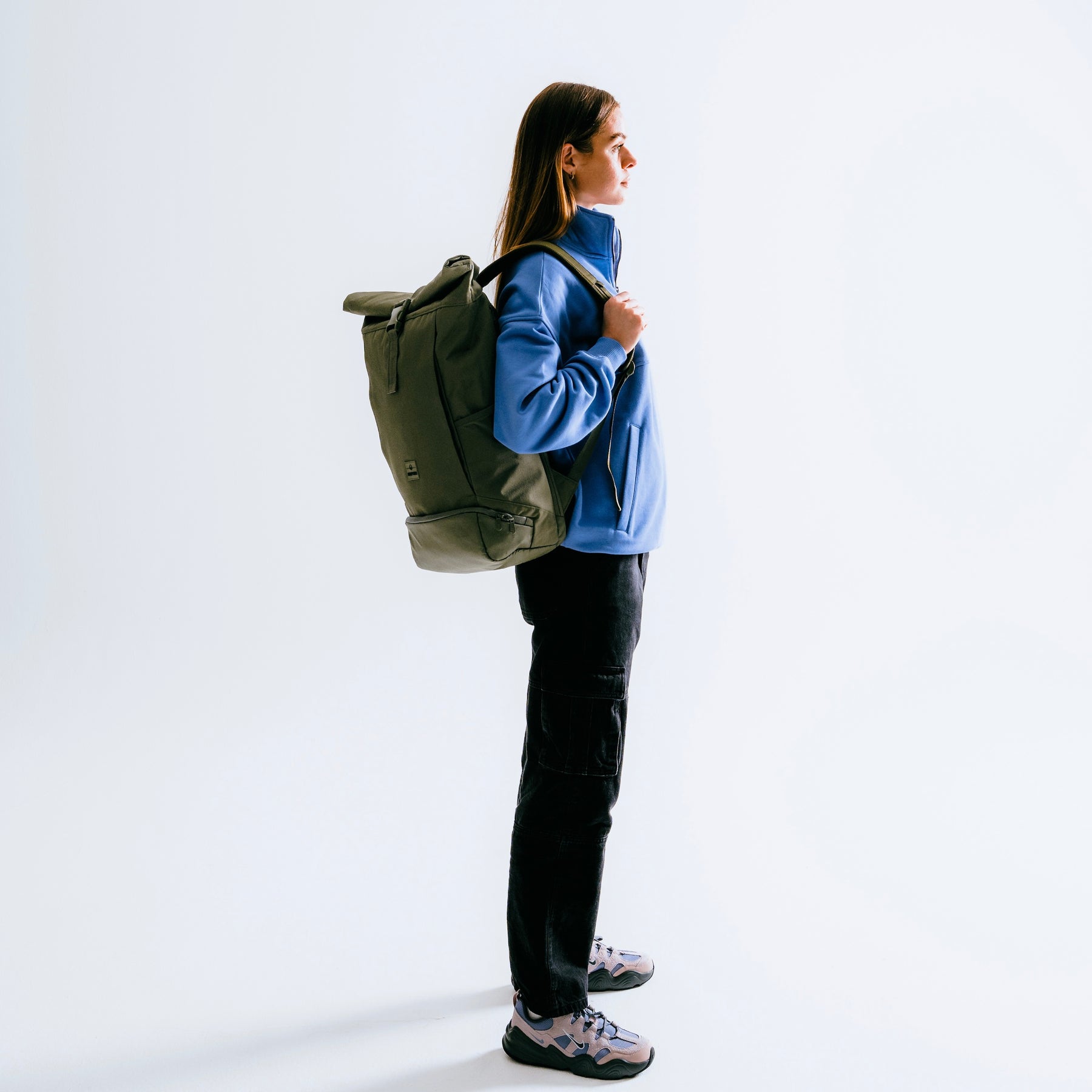 Frau im Profil mit olivgrünem Rolltop Rucksack 'Allen Large', blauem Oberteil, schwarzer Hose und Sportschuhen