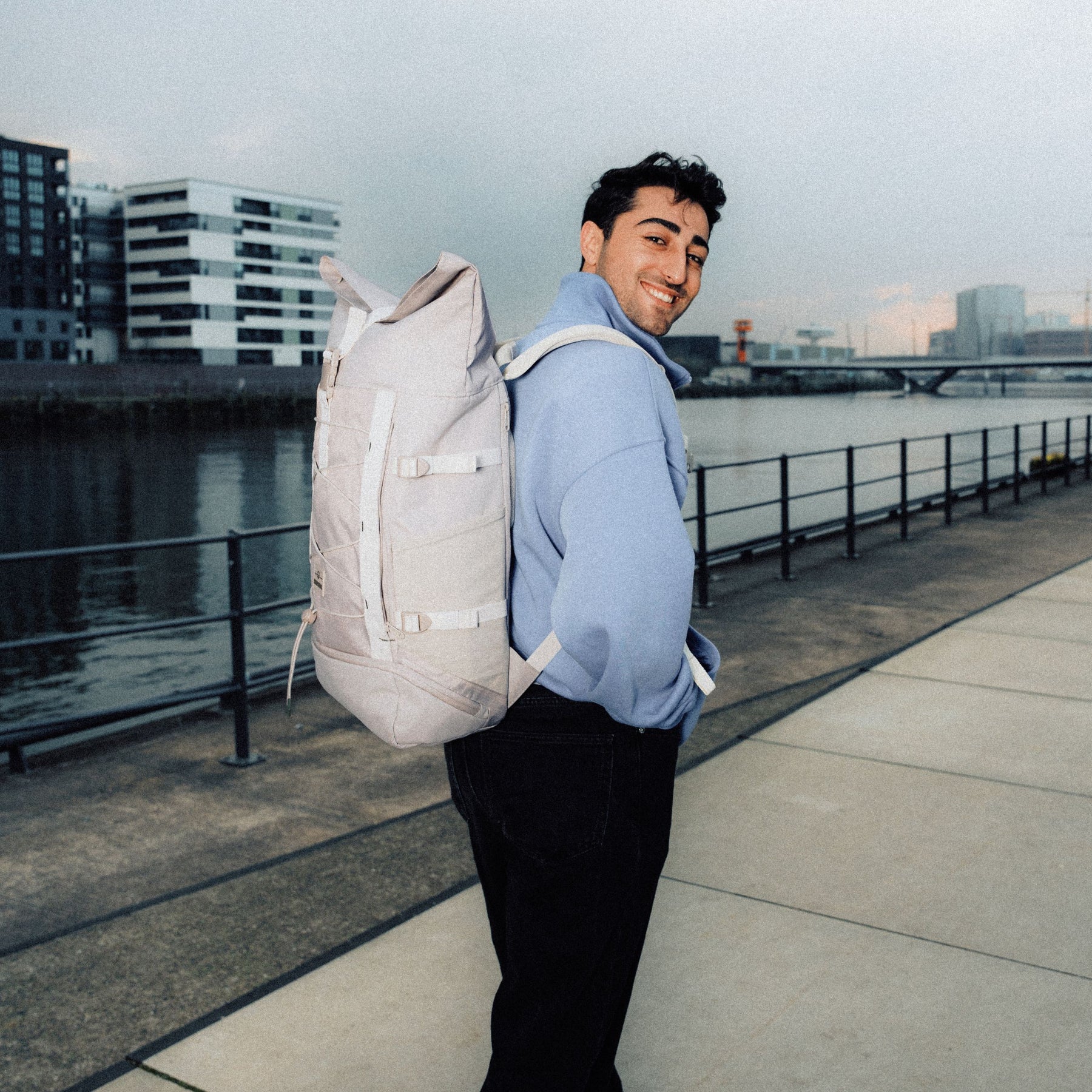 Mann trägt beigen Rolltop Rucksack 'Allen Large Move' und hellblauen Pullover am Wasser in urbaner Umgebung