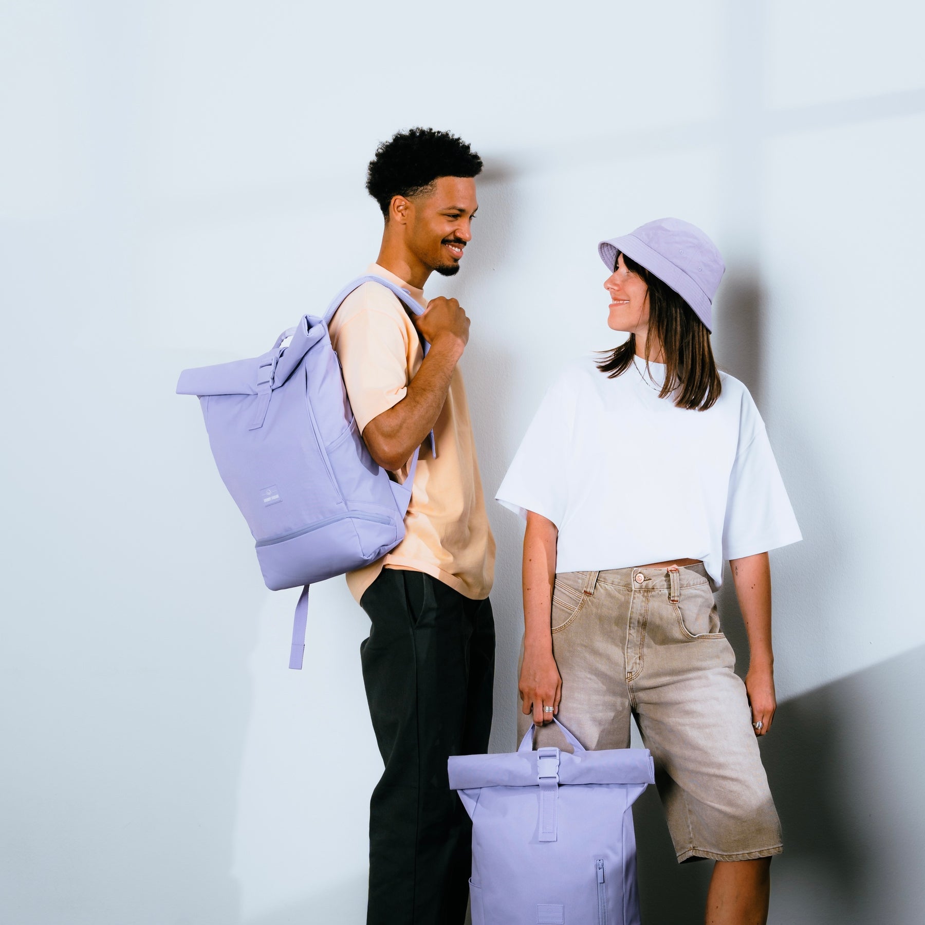 Mann trägt hellvioletten Rolltop-Rucksack 'Allen Medium', Frau hält passenden Rucksack, beide in lässiger urbaner Kleidung