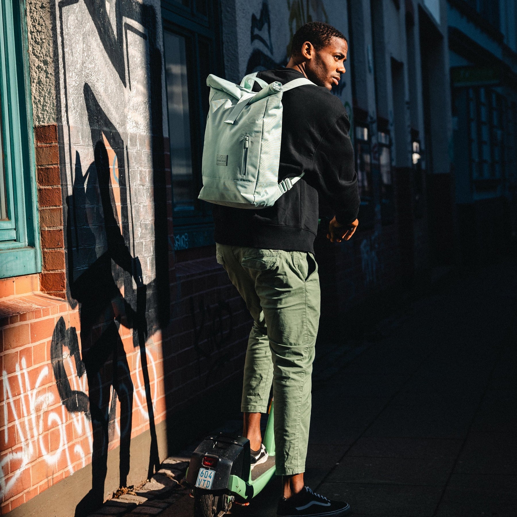 Mann trägt mintgrünen Rolltop-Rucksack 'Robin Medium' in urbaner Umgebung auf grünem E-Scooter