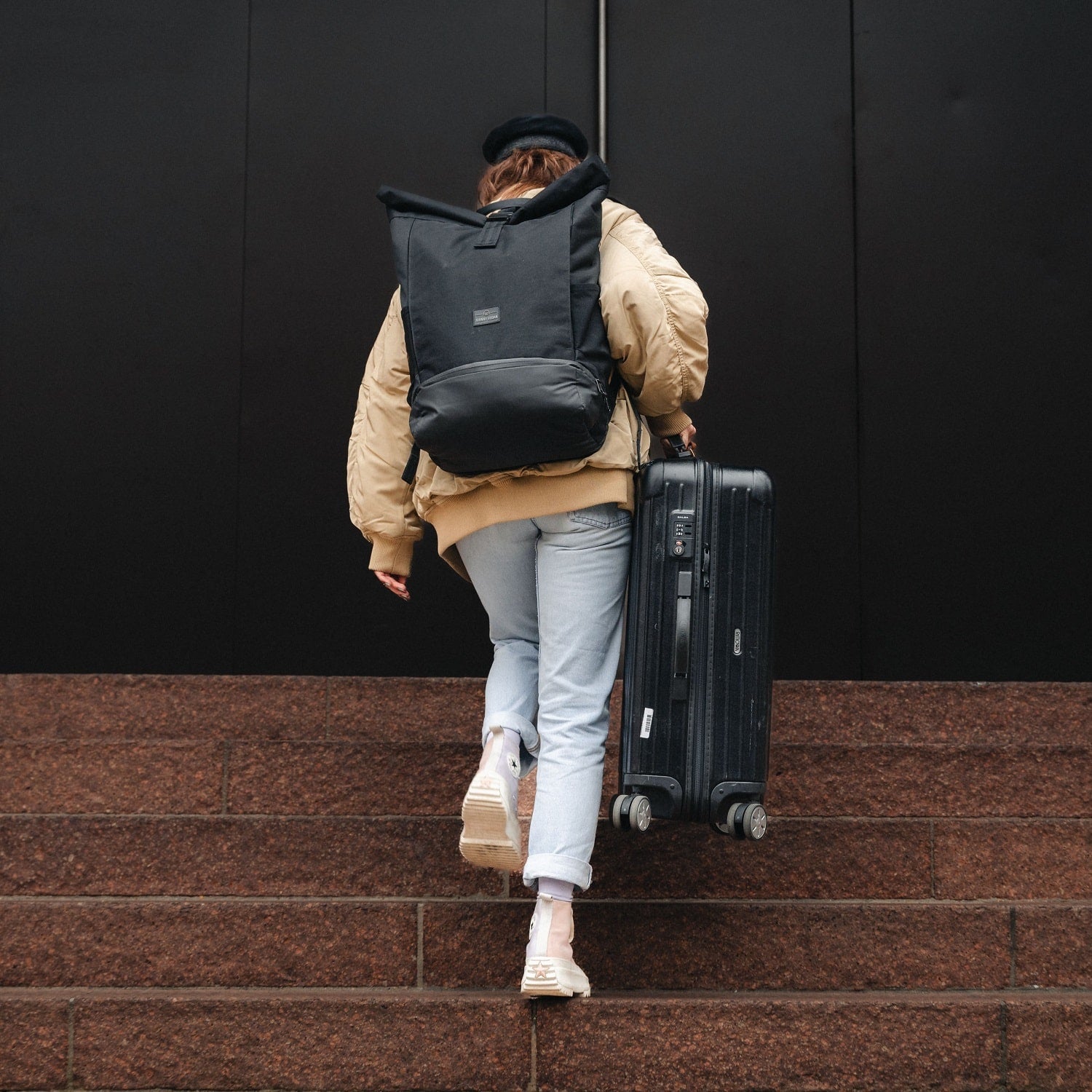 Person trägt schwarzen Rolltop Rucksack Allen XL und zieht schwarzen Koffer eine Treppe hinauf