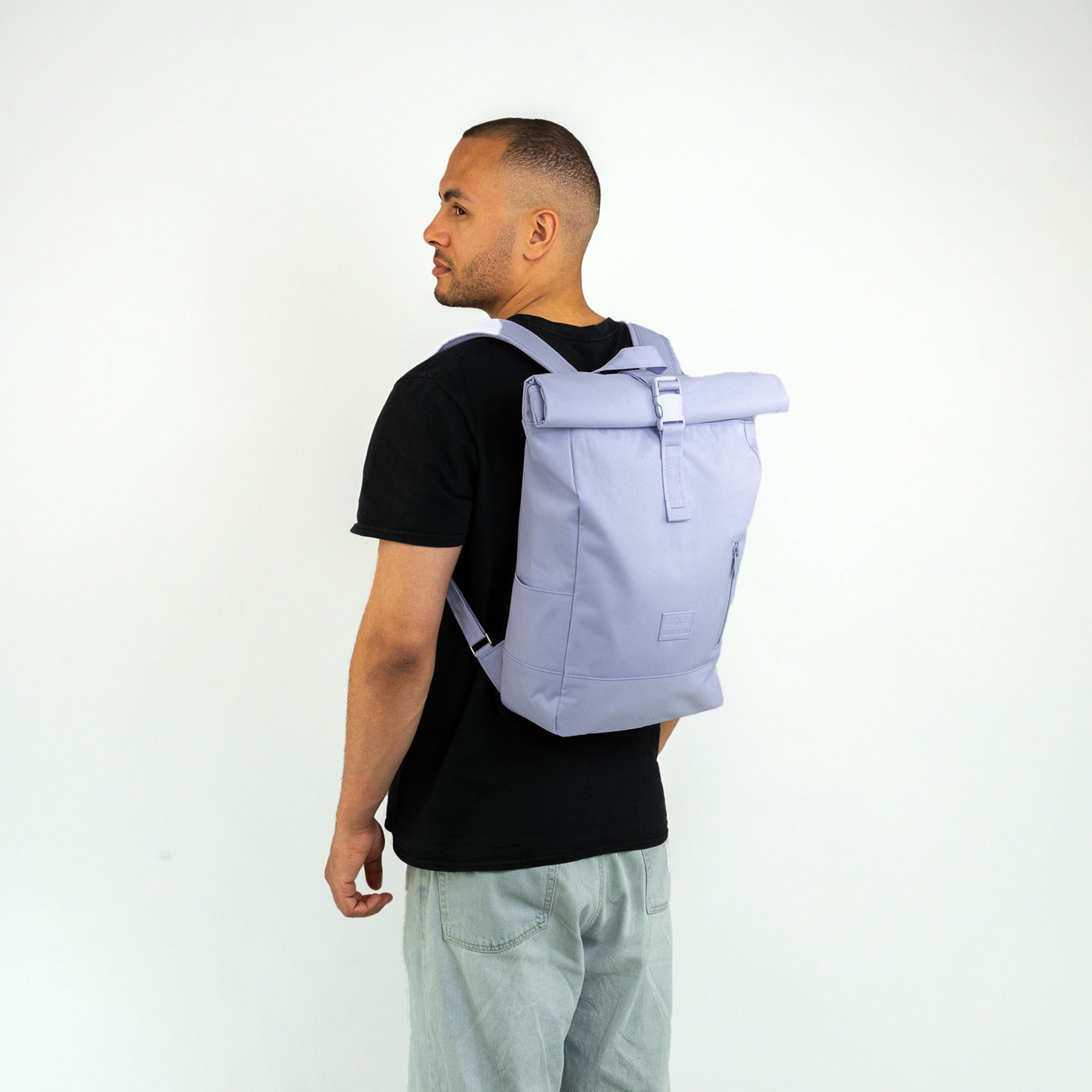 Mann trägt Johnny Urban Rolltop Rucksack 'Robin Medium' in flieder-lila mit schwarzem T-Shirt und hellen Hosen