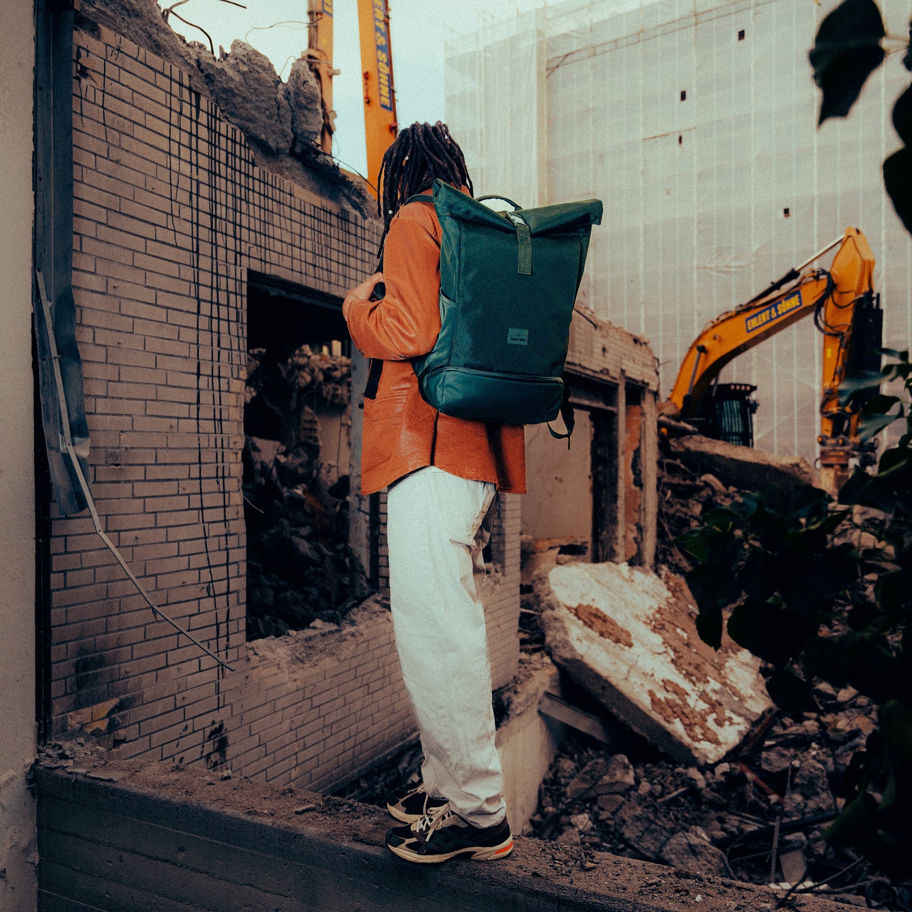 Mann mit dunkelgrünem Rolltop-Rucksack 'Allen Large' von Johnny Urban vor einer Baustelle mit Bagger