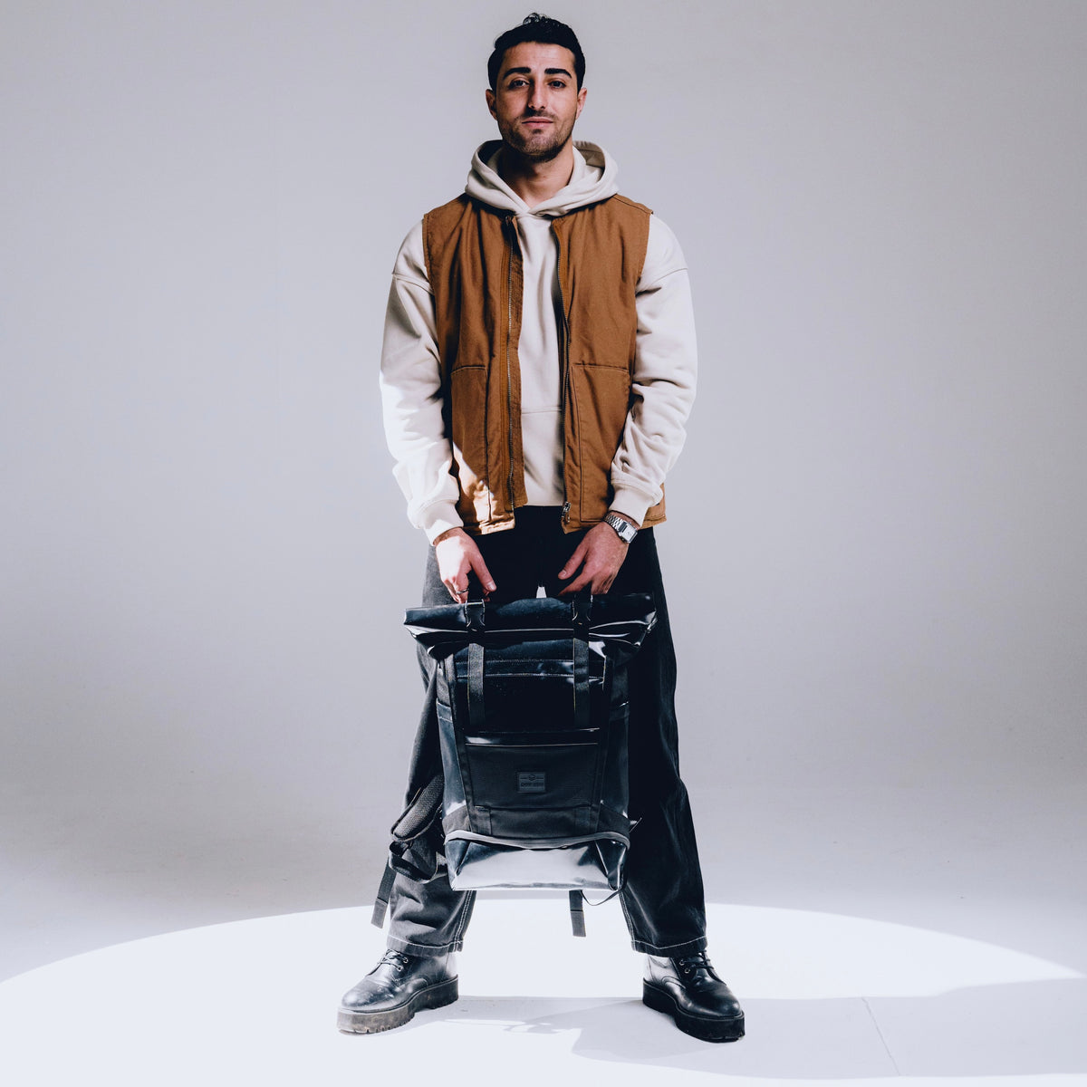 Mann mit beigem Kapuzenpullover und brauner Weste hält schwarzen Rolltop Rucksack 'Ashton' von Johnny Urban
