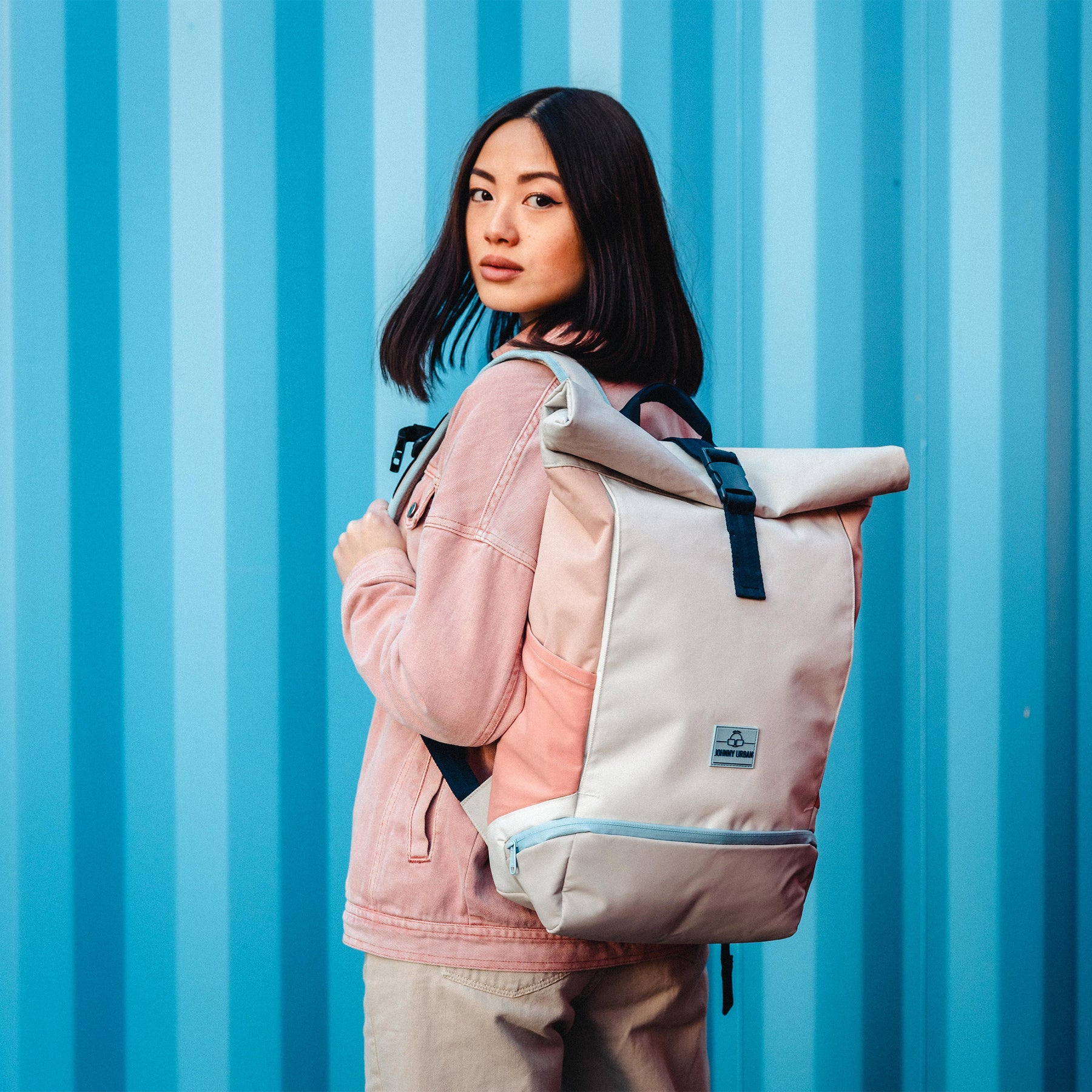 Frau trägt Rolltop Rucksack 'Allen Medium' in Beige-Rose mit schwarzem Verschluss, vor blauem Hintergrund