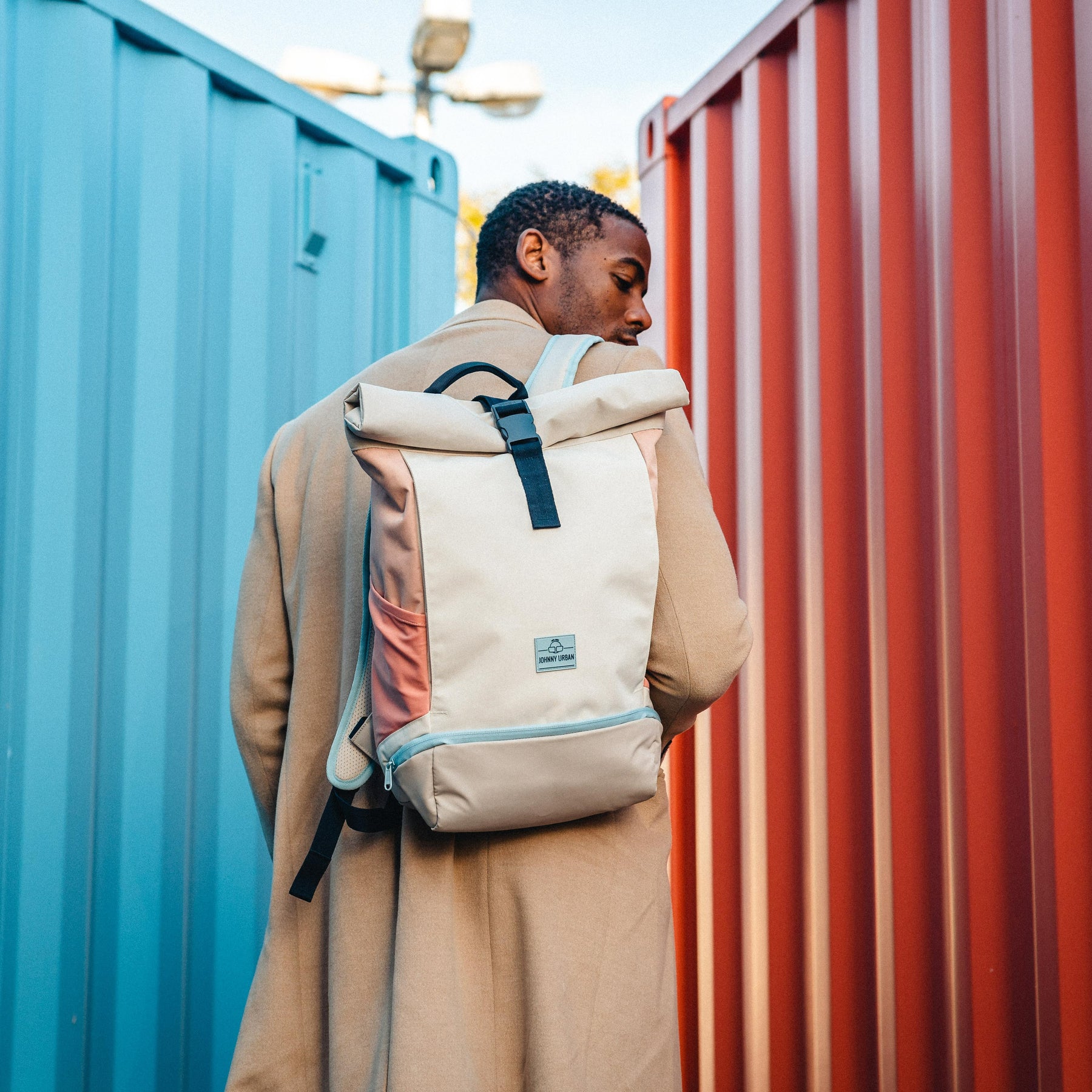 Mann trägt Johnny Urban Rolltop Rucksack 'Allen Medium' in Beige-Rose vor blauen und roten Containern