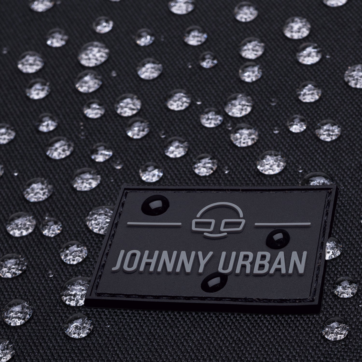 Nahaufnahme der wasserabweisenden schwarzen Oberfläche des Rolltop Rucksacks 'Allen Medium' mit JOHNNY URBAN Logo und Wassertropfen