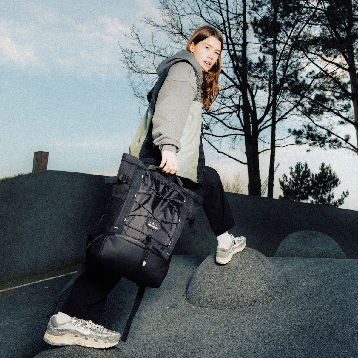 Frau trägt schwarzen Rucksack 'Harper' von Johnny Urban, sitzt auf einer grauen Steinstruktur im Freien