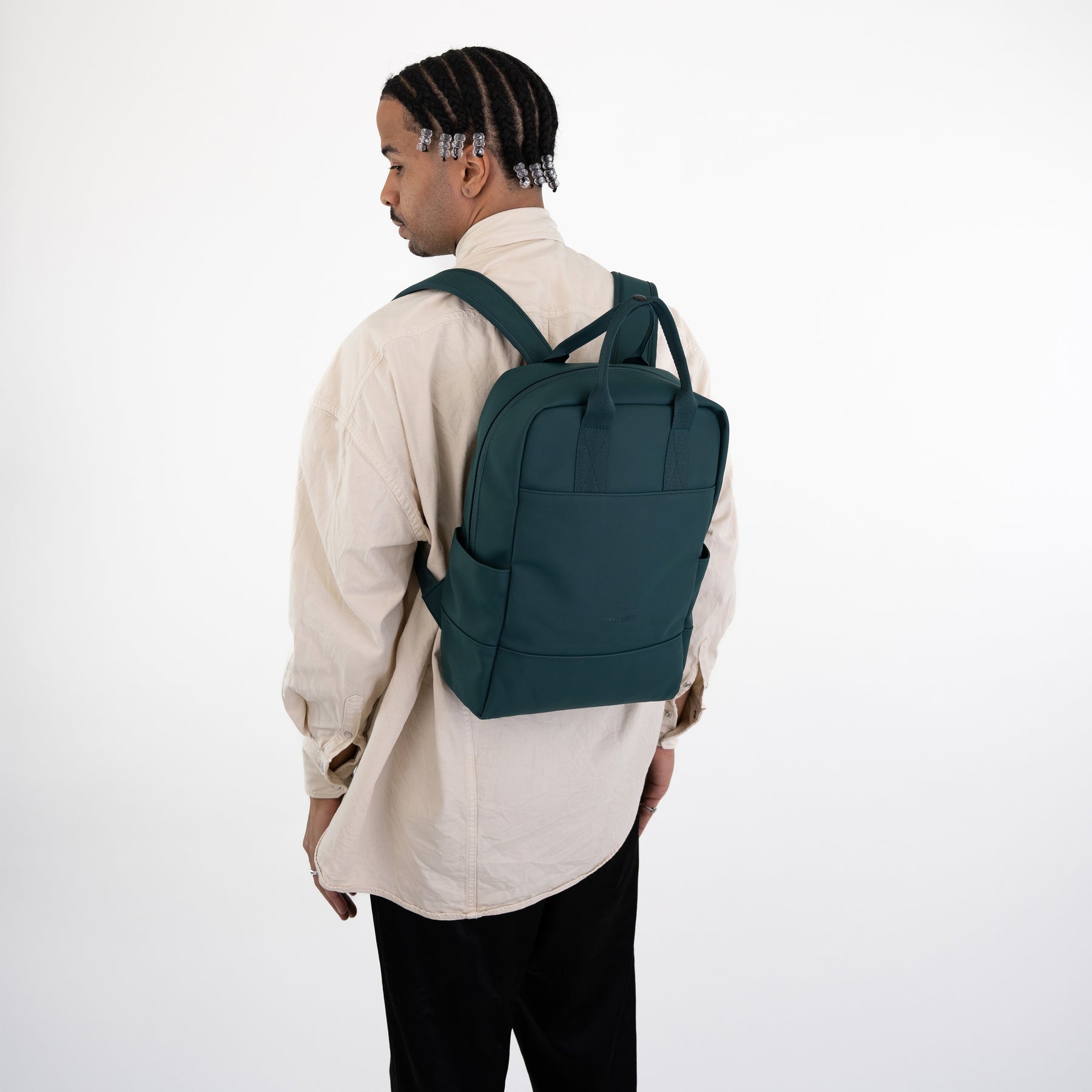 Mann mit Zöpfen trägt grünen Rucksack 'Hailey' aus Stoff, beige Hemd, schwarze Hose vor weißem Hintergrund