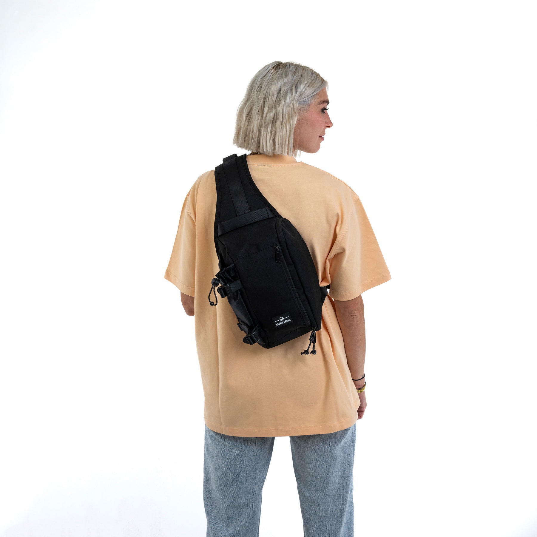 Frau mit kurzem blondem Haar trägt schwarze Johnny Urban Sling Bag 'Charlie' über beigem T-Shirt und blauer Jeans