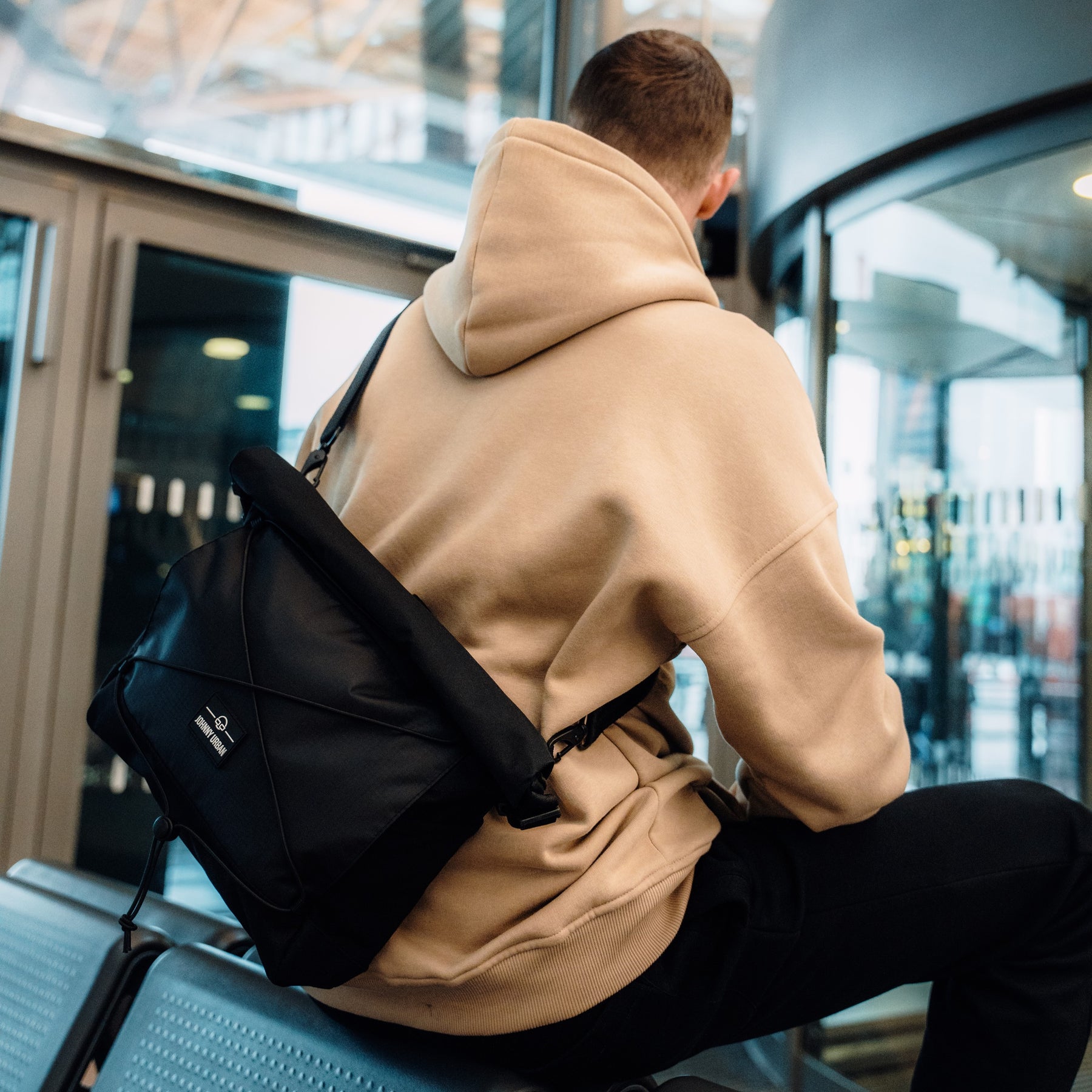 Schwarze Johnny Urban Umhängetasche 'Dylan' aus robustem Material, getragen von einem Mann im beigen Hoodie in moderner Umgebung