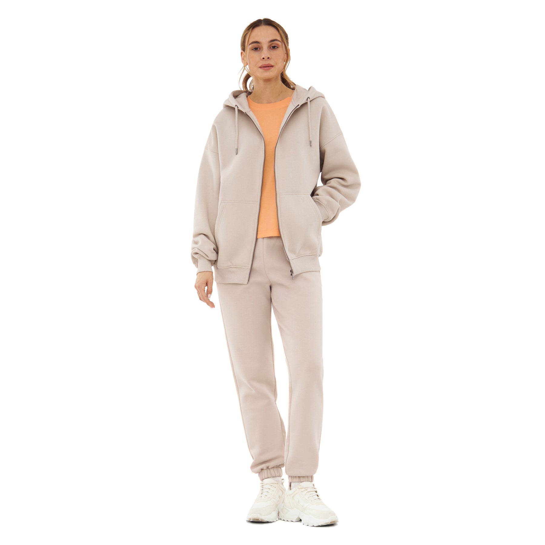 Frau trägt beige Oversized Zip Hoodie 'Caleb' mit Kapuze und bequeme Jogginghose, darüber orange T-Shirt