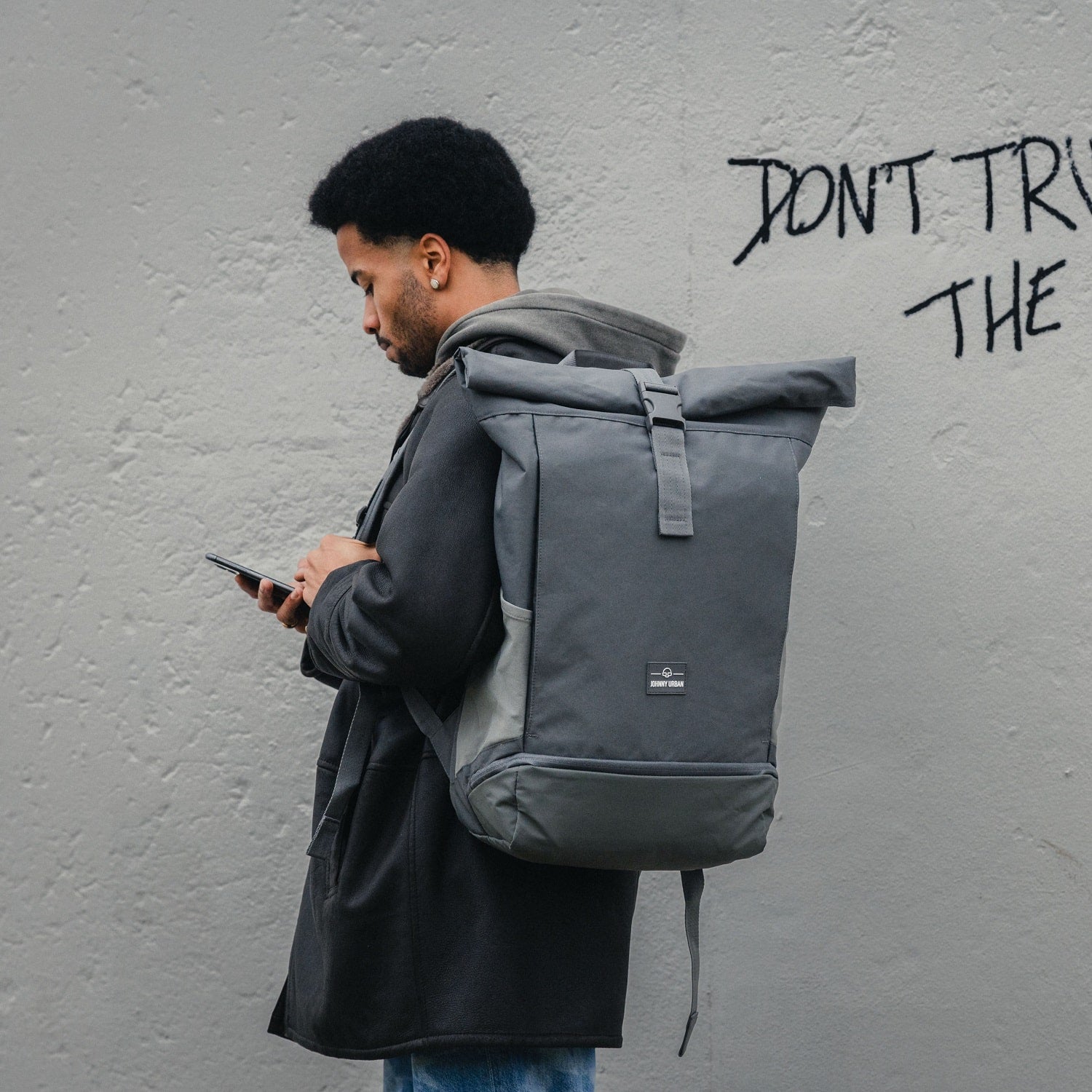 Mann trägt grauen Rolltop Rucksack Allen XL von Johnny Urban und nutzt Handy vor Wand mit Text