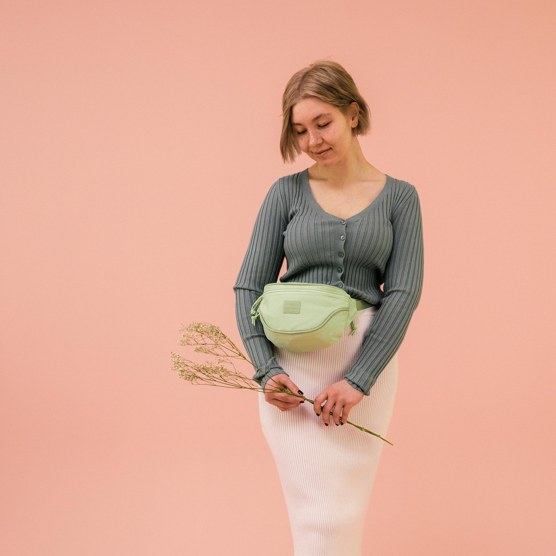 Frau mit grüner Bauchtasche ‚Ben‘ von Johnny Urban, grauer Strickjacke und weißem Rock vor rosa Hintergrund
