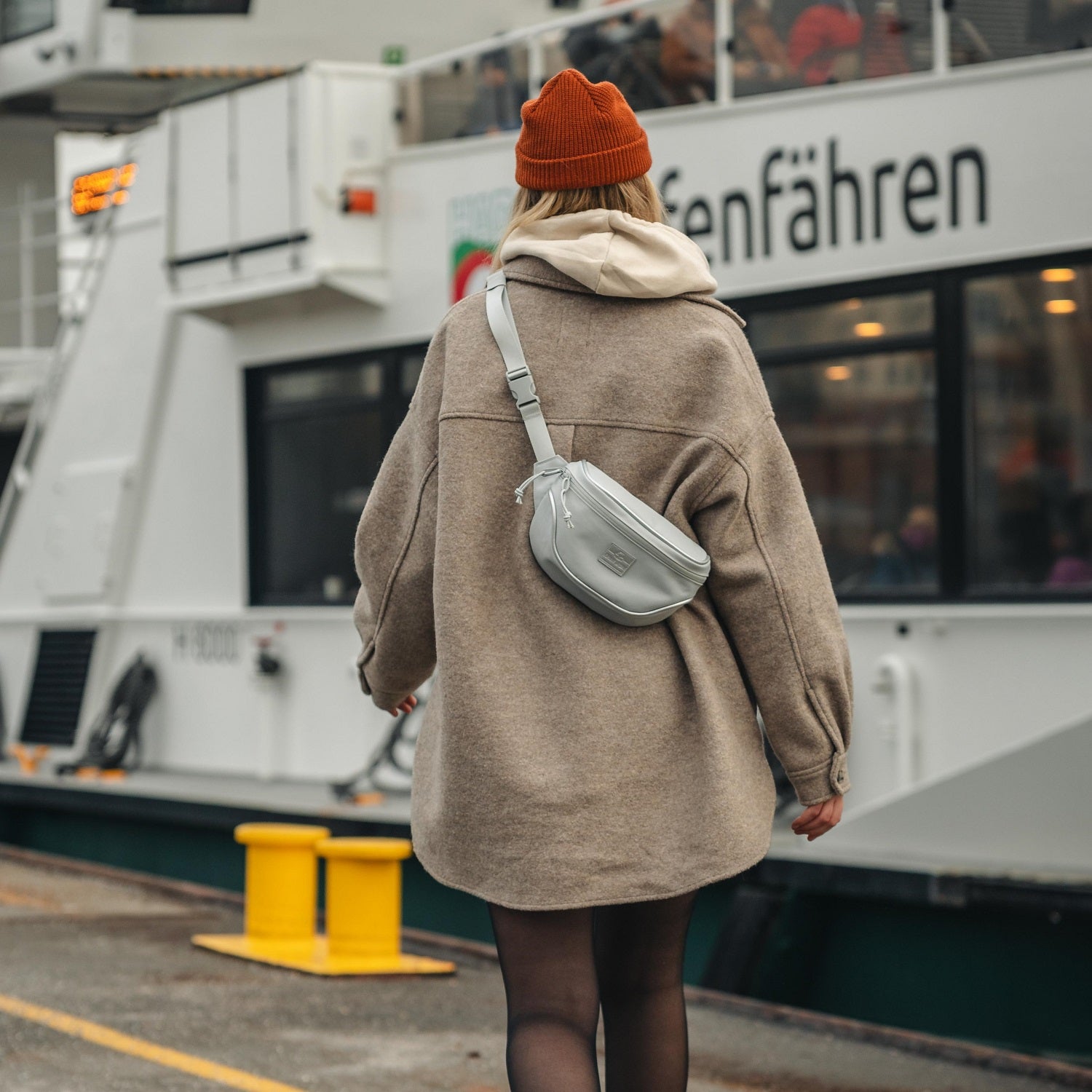 Dame trägt graue Johnny Urban Bauchtasche 'Ben' quer über dem Rücken, brauner Mantel, roter Strickmütze, im urbanen Hafenbereich