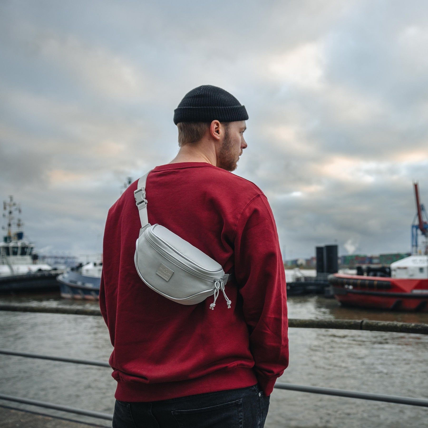 Mann trägt graue Bauchtasche 'Ben' von Johnny Urban über rotem Pullover am Hafen
