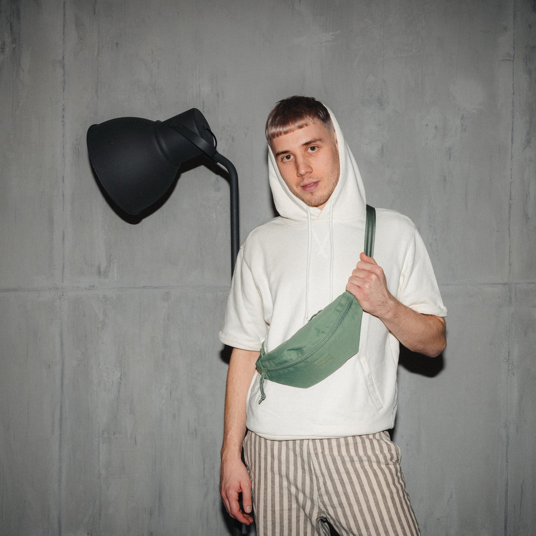 Mann trägt grüne Bauchtasche 'Erik Small' von Johnny Urban über der Schulter, weiße Kapuzenjacke, gestreifte Hose