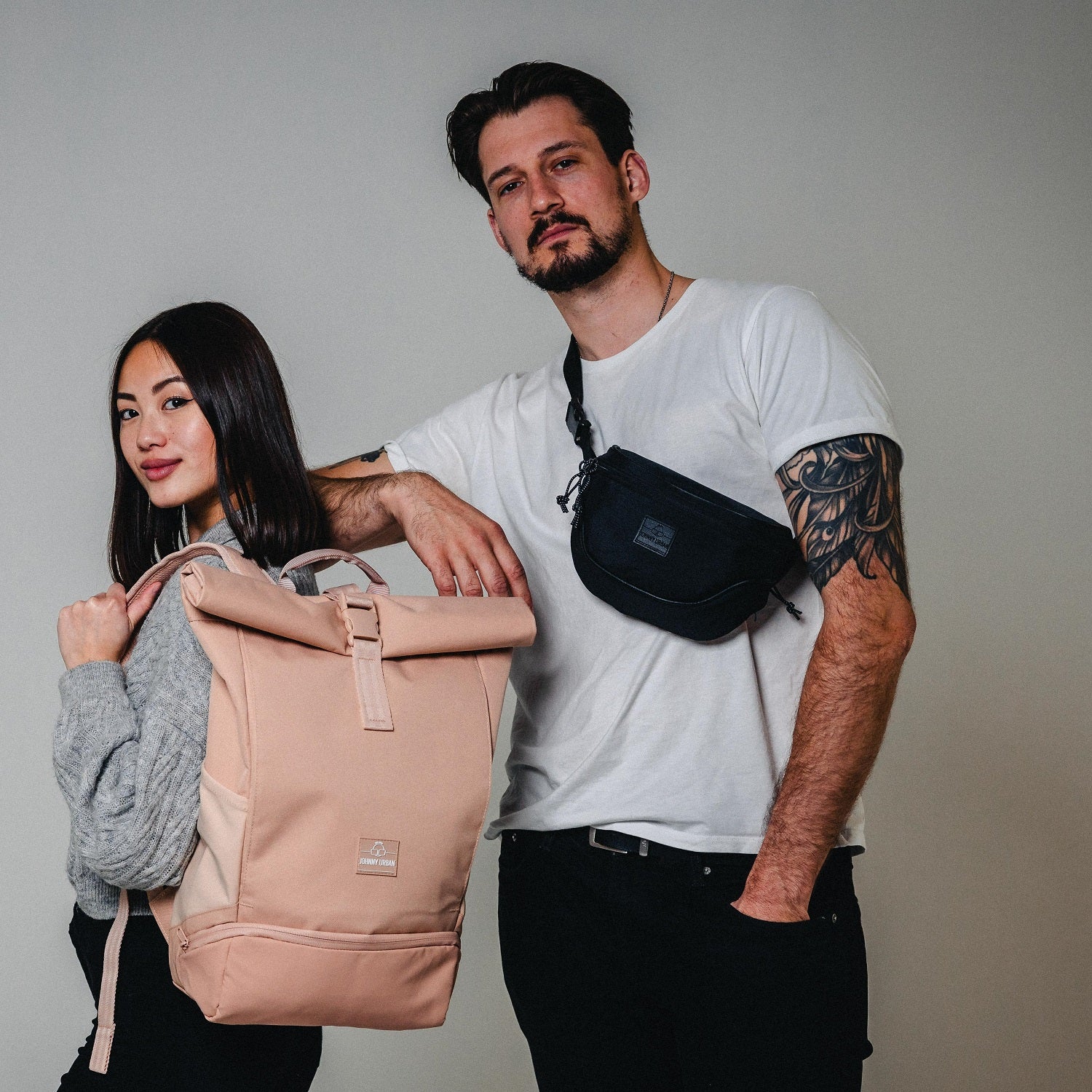 Mann trägt schwarze Bauchtasche 'Ben' von Johnny Urban quer über die Brust, Frau hält beigen Rucksack mit Rollverschluss
