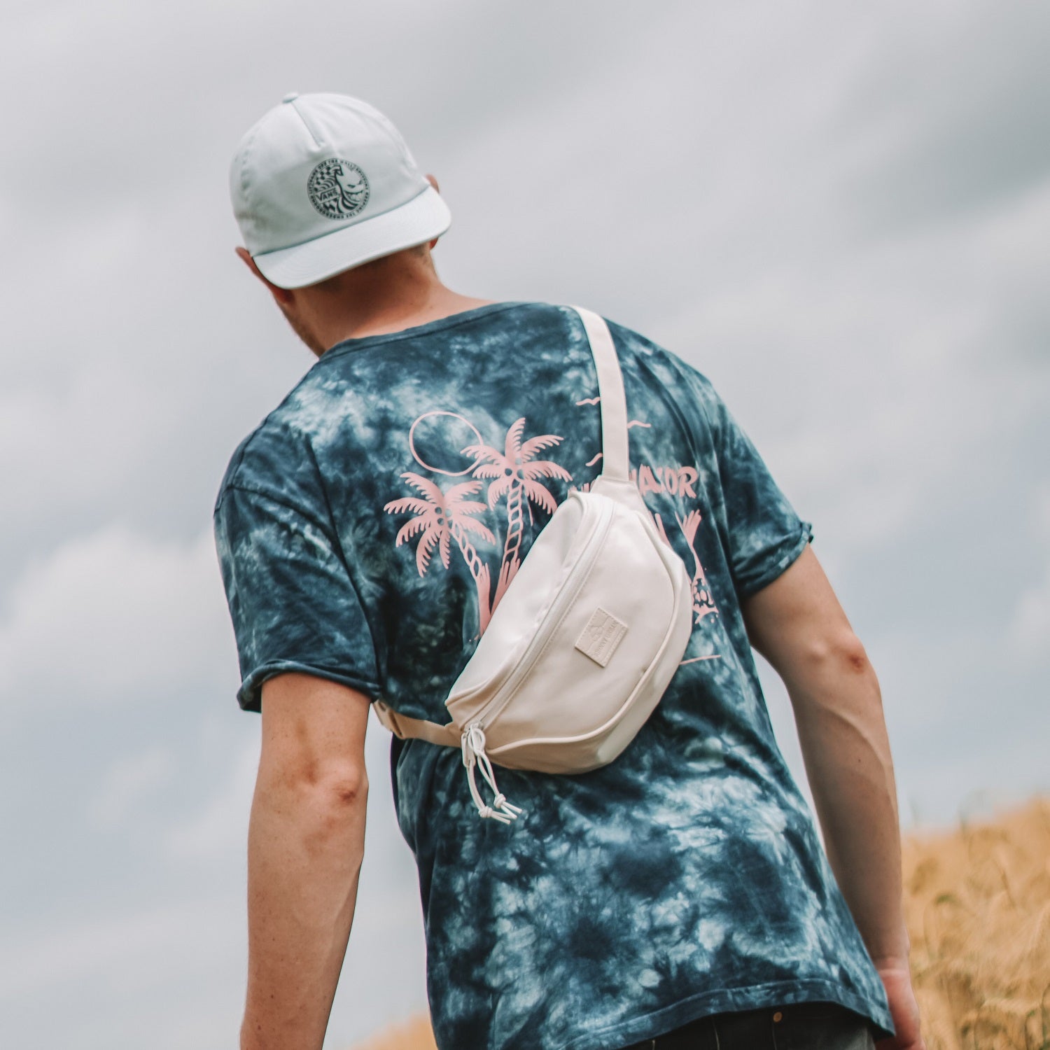 Mann von hinten trägt weiße Bauchtasche 'Ben' von Johnny Urban quer über den Rücken, dazu blau-schwarzes Batik-T-Shirt mit pinken Palmen