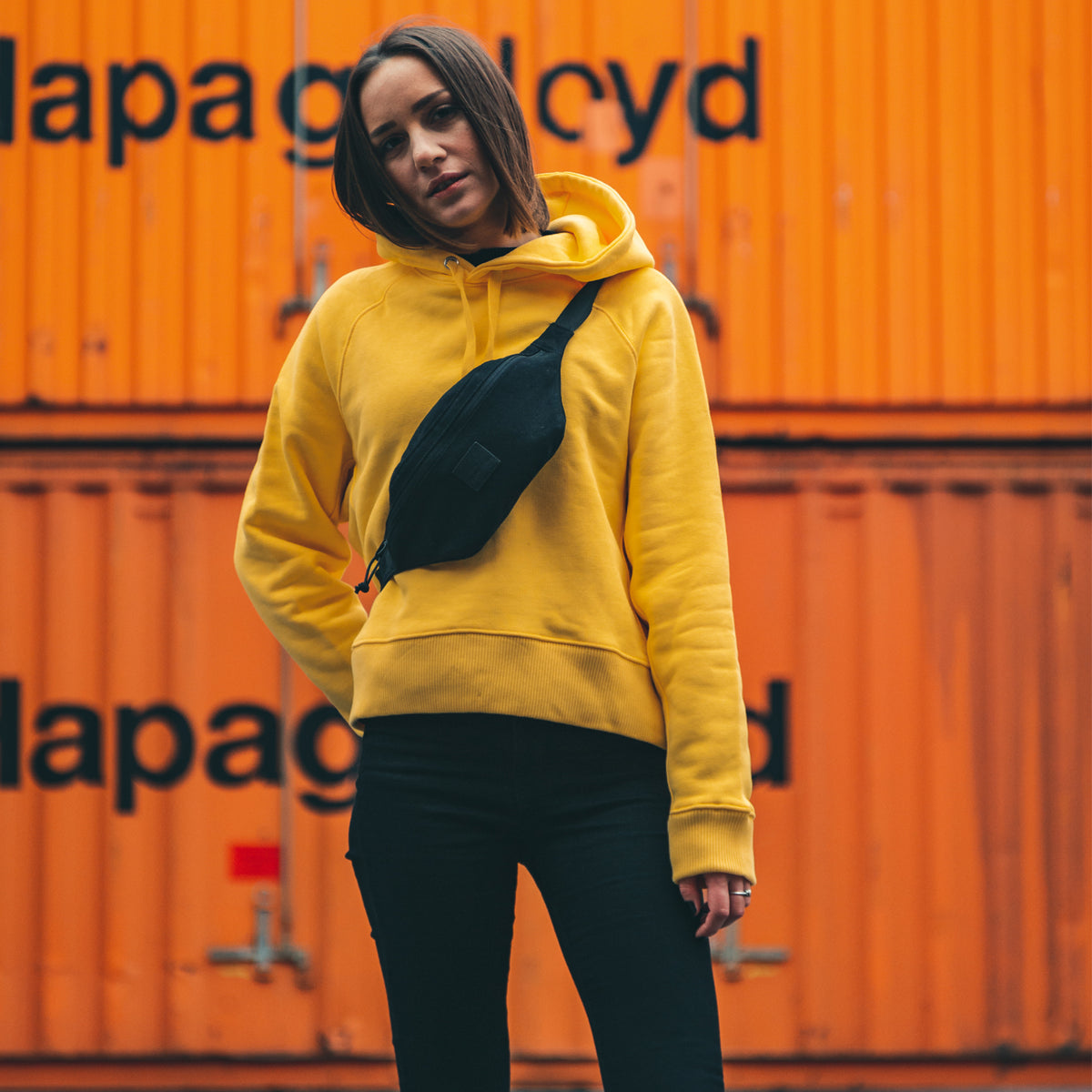 Frau trägt gelben Hoodie und schwarze Bauchtasche ‚Toni‘ quer über der Brust vor orangefarbenen Containern