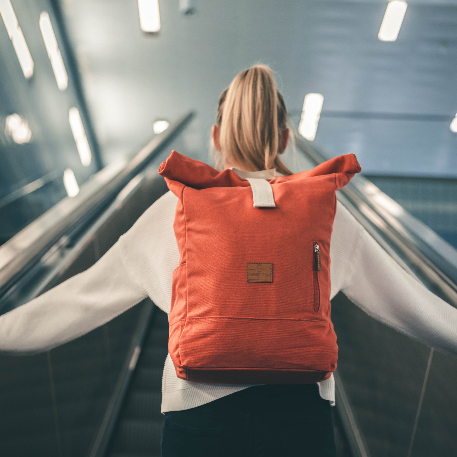 Rückenansicht einer Frau, die einen roten Rolltop Rucksack ‚Adam Large‘ trägt, stehend vor einer Rolltreppe