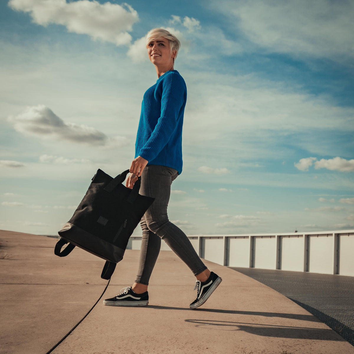 Frau mit kurzem blondem Haar trägt schwarzen Canvas-Rucksack 'Sam' in der Hand, trägt blauen Pullover und graue Jeans