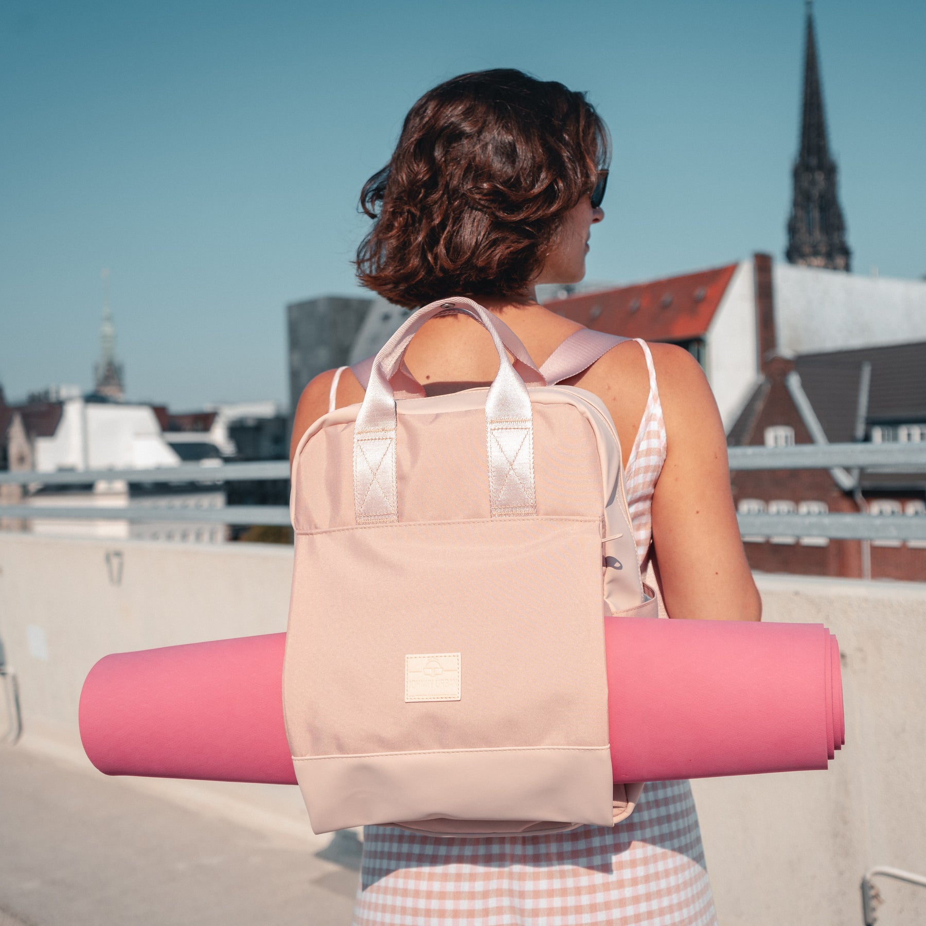 Rucksack 'Jona Large' in Rosa mit integriertem Tragegurt hält eine Yogamatte, getragen von Frau in ärmellosem Kleid