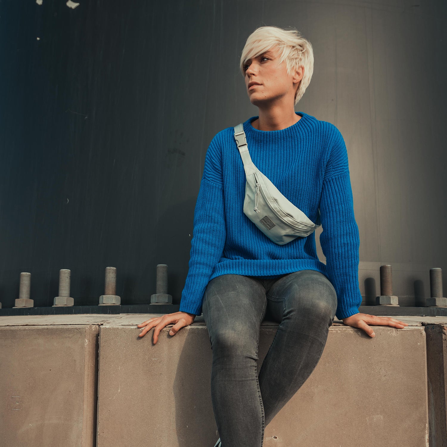 Frau mit kurzem blondem Haar trägt blaue Strickjacke, graue Jeans und graue Bauchtasche 'Erik Small' quer über die Brust