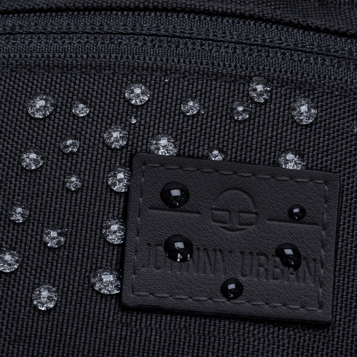 Nahe Aufnahme einer wasserabweisenden schwarzen Bauchtasche 'Erik Small' mit Wassertropfen auf dem Stoff und dem Johnny Urban Label