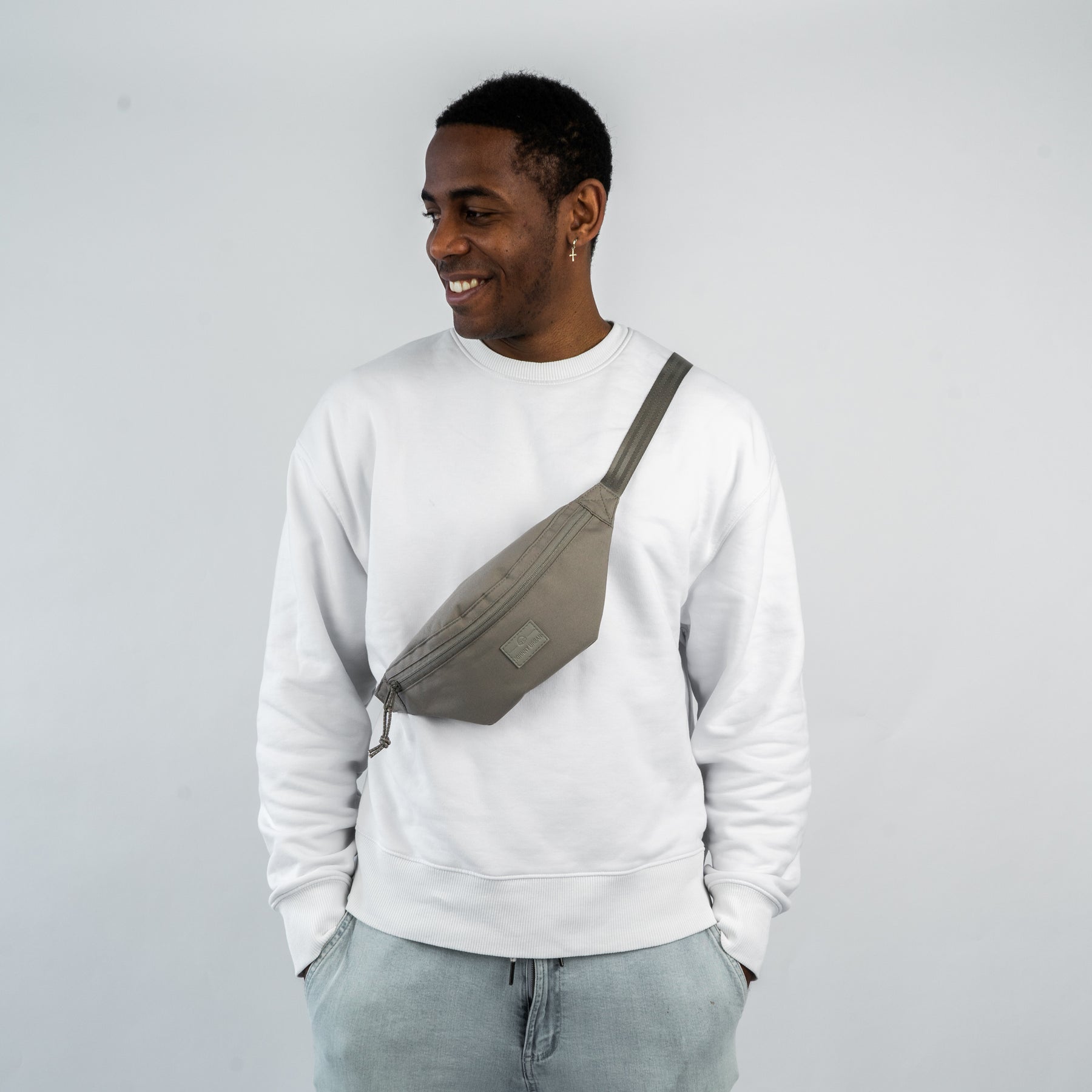 Mann trägt die Johnny Urban Bauchtasche 'Erik Small' in Grau über der Brust, kombiniert mit weißem Sweatshirt und hellblauen Jeans